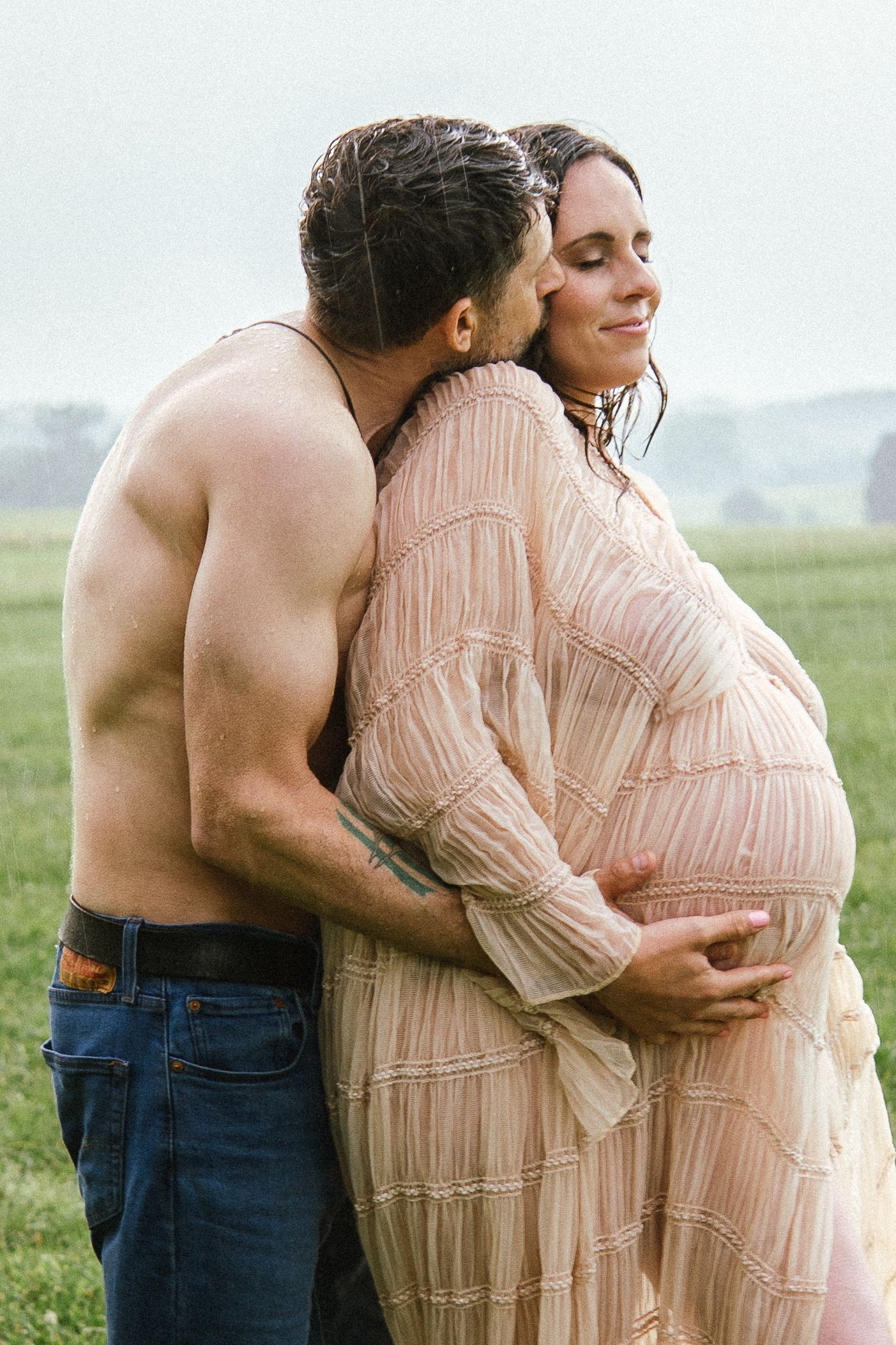 A couple embracing in a grassy field, she wearing a flowing peach-colored dress, he shirtless in jeans.