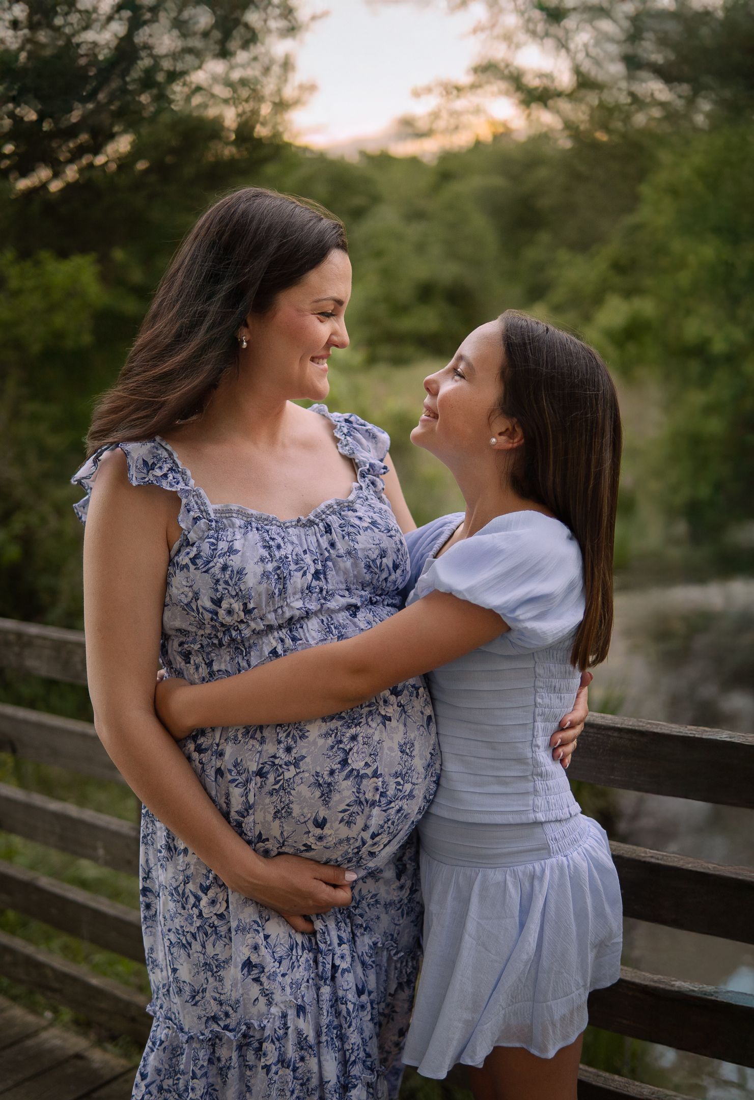 A pregnant woman in a floral dress shares a tender moment with her daughter against a sunset backdrop by a wooden fence.