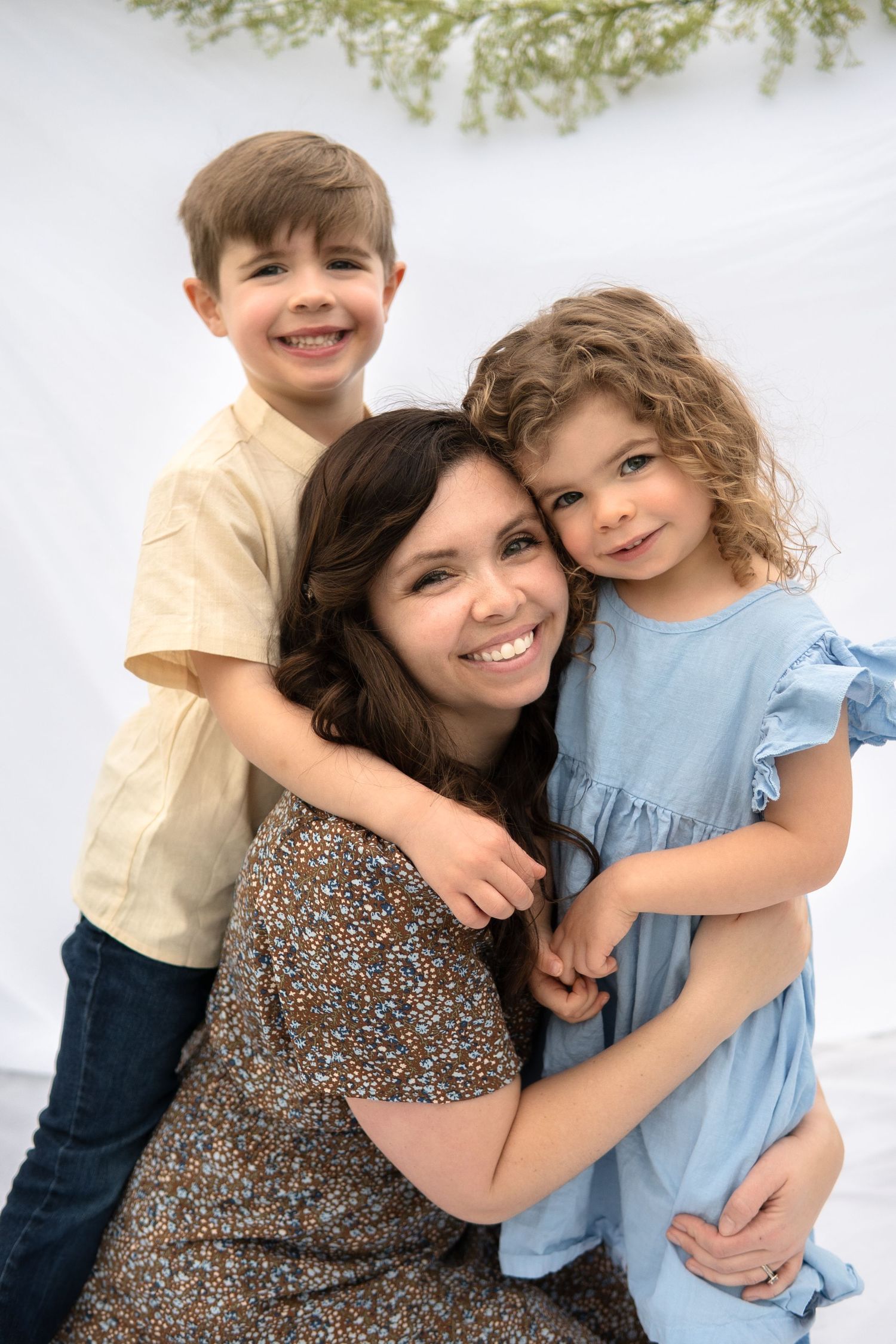 Loving family portrait with mother in floral dress hugging children in casual attire against white background.