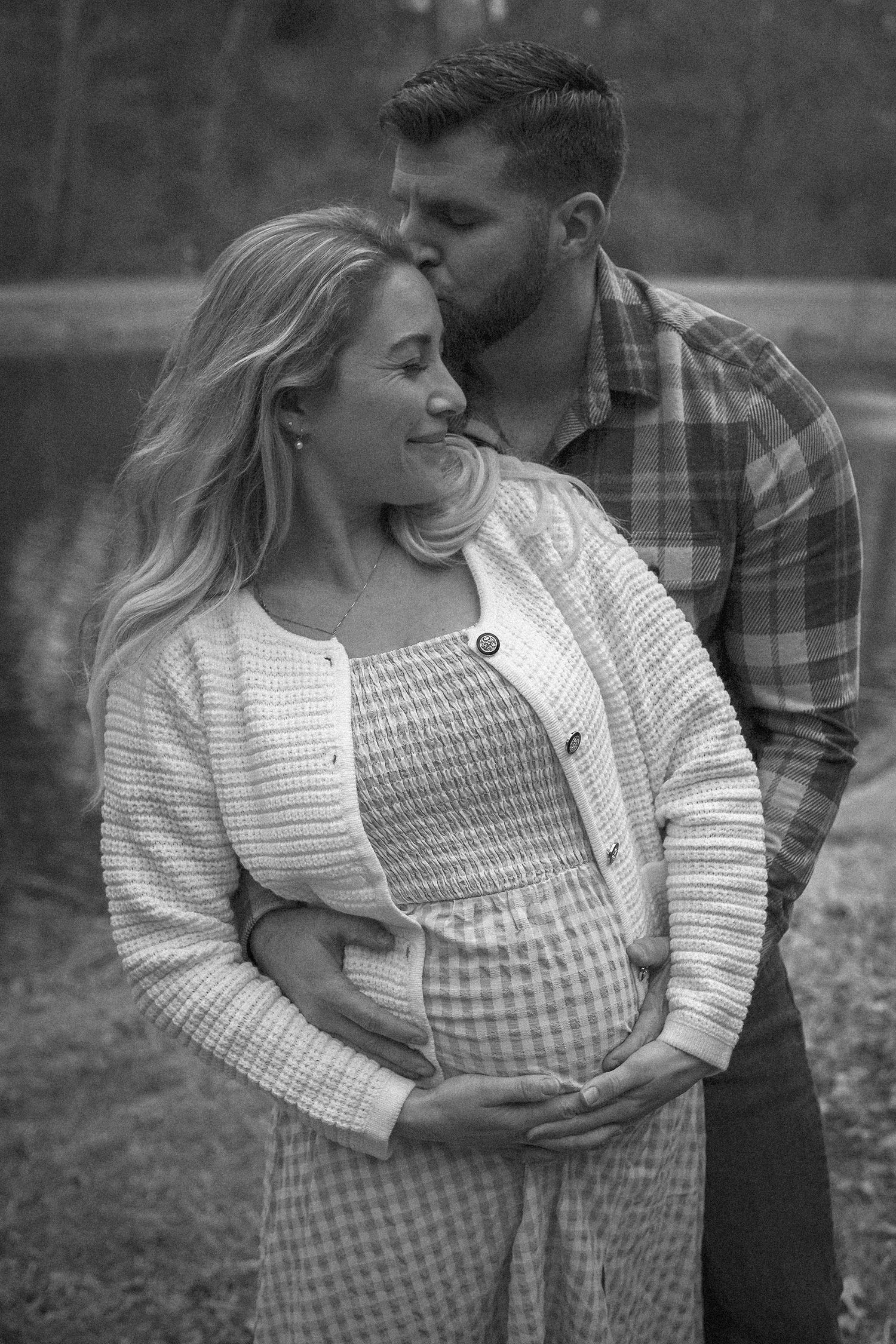 A tender black and white maternity photo shows a couple embracing outdoors, with her wearing a checkered dress and sweater.