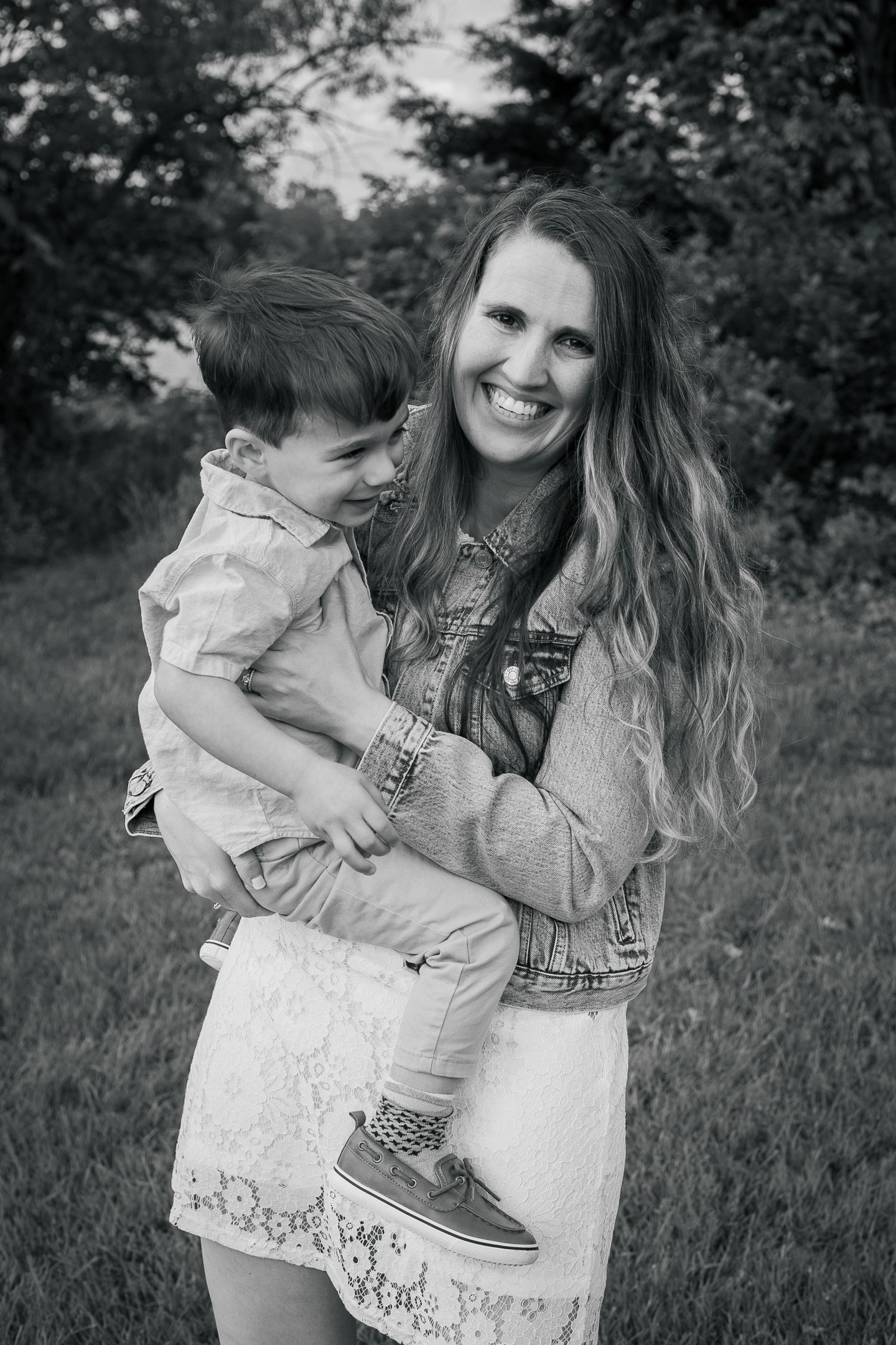 A candid black and white portrait of a woman in a denim jacket and white dress holding a small child outdoors.