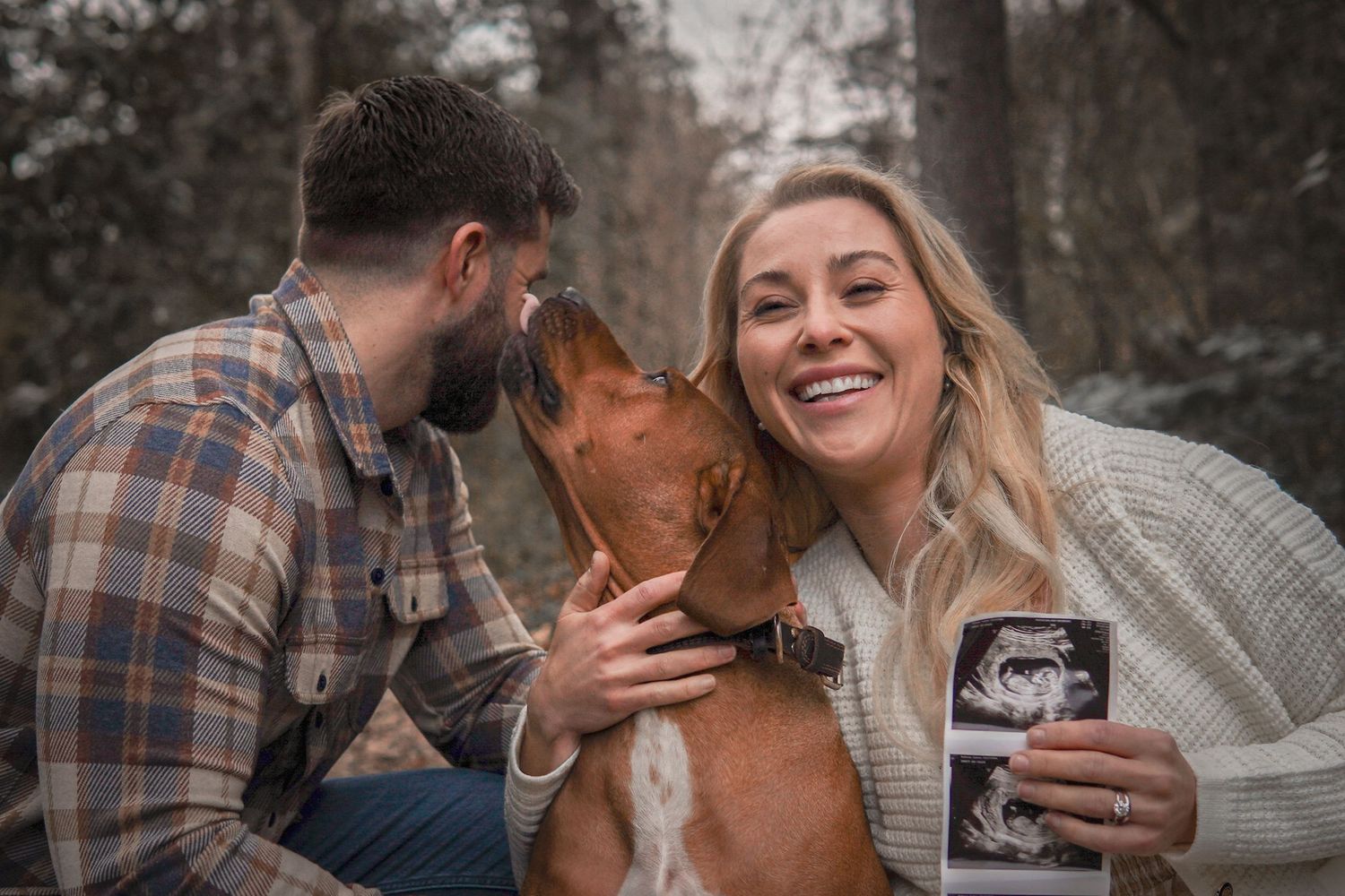 A couple shares a joyful moment outdoors with their brown dog while holding an ultrasound photo.