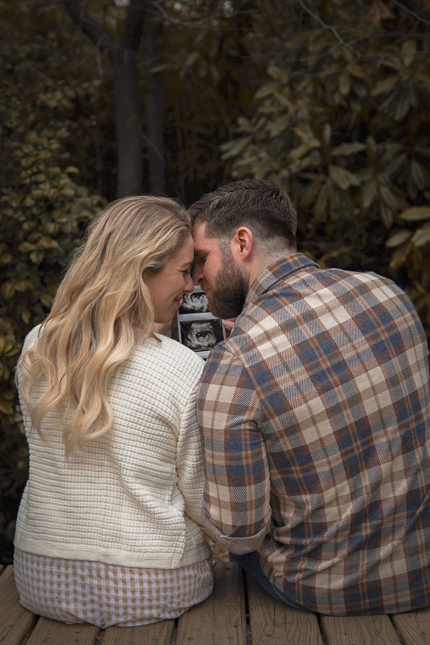 A couple sits intimately on wooden steps sharing an ultrasound photo in an autumn woodland setting.