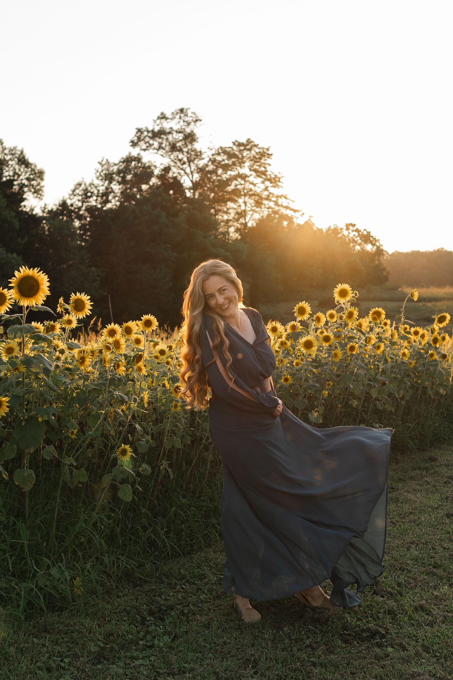 Long flowing navy dress twirls in a sunflower field at golden hour sunset with sunlight streaming through trees.
