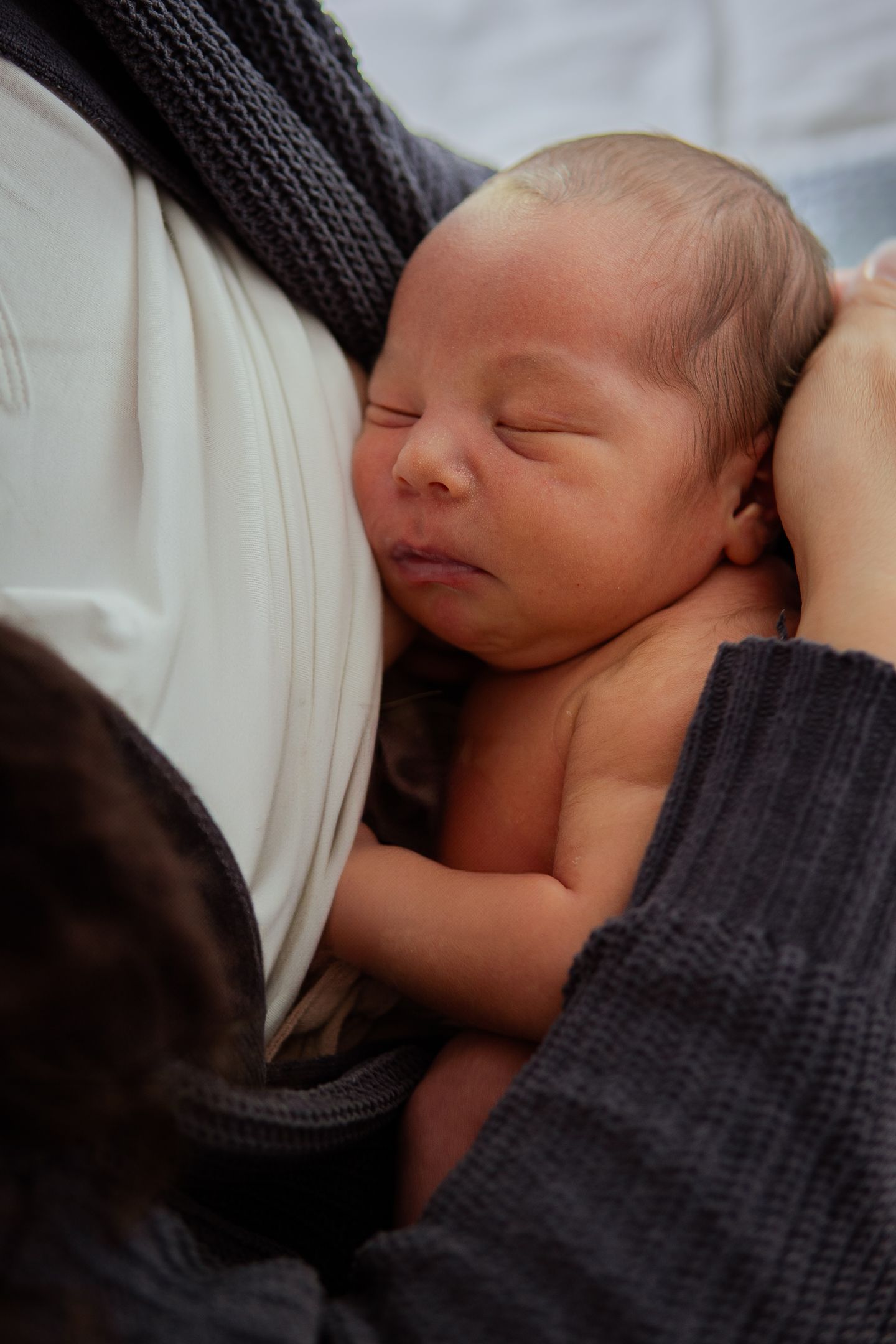 Peaceful sleeping newborn rests against parent wearing white shirt and dark sweater.