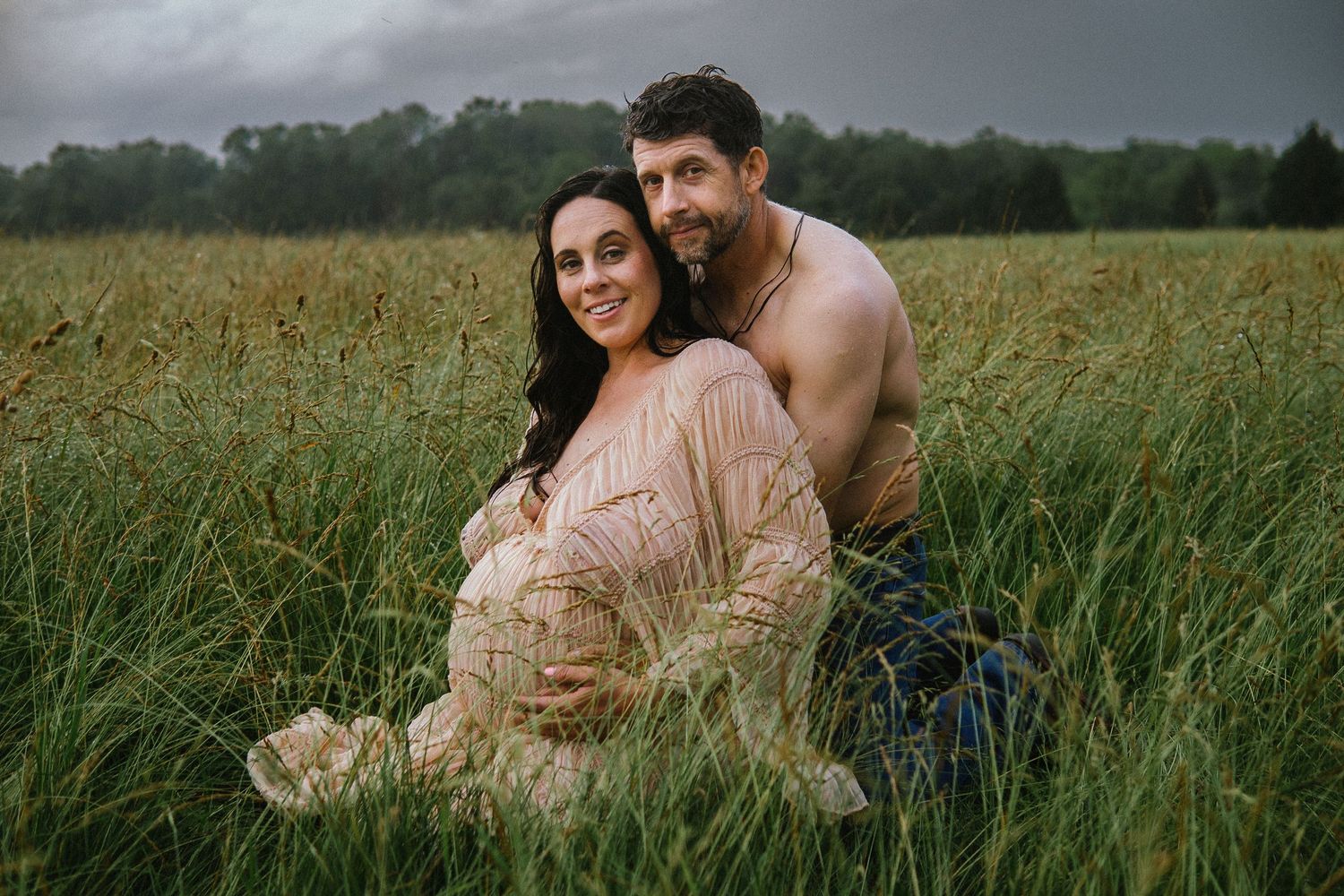 A couple shares an intimate moment in a rustic field during a maternity photoshoot at sunset.