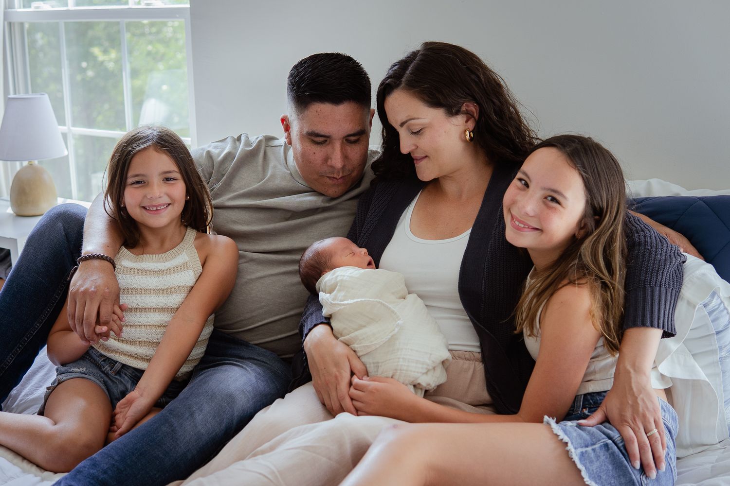 Family sitting together on bed looking at newborn baby wrapped in white blanket.