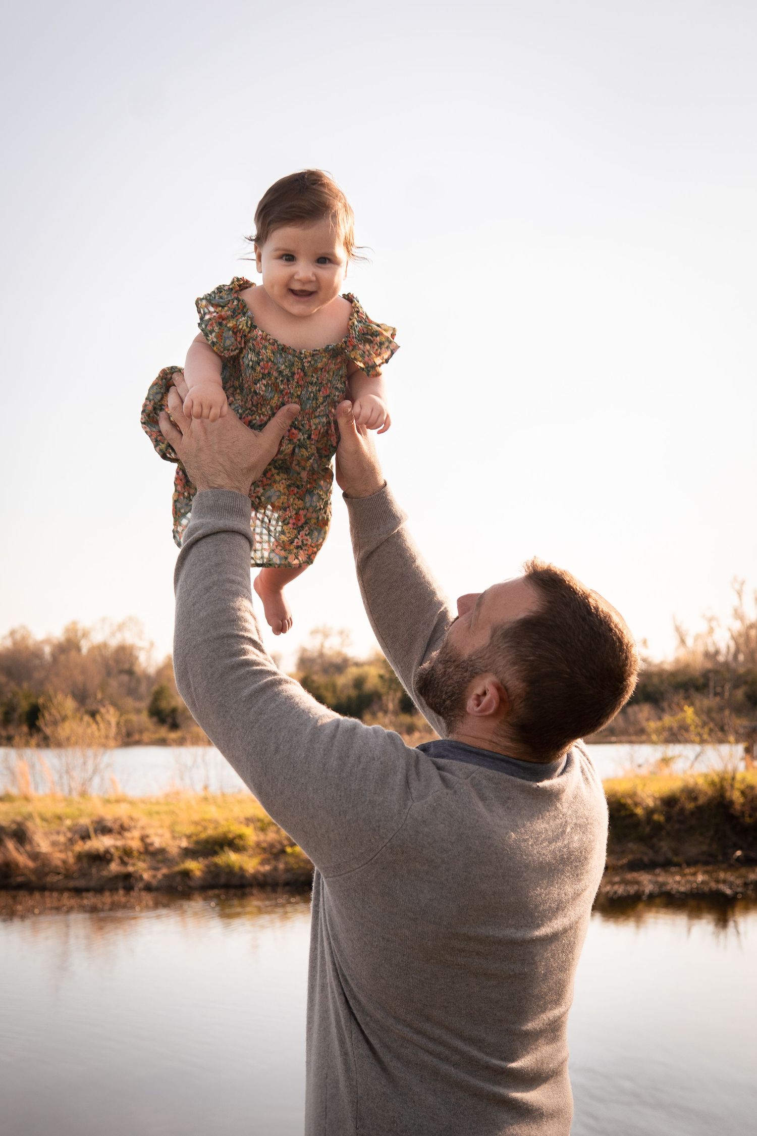 A joyful outdoor moment by the water as a person in gray lifts a laughing baby wearing a floral dress at sunset.