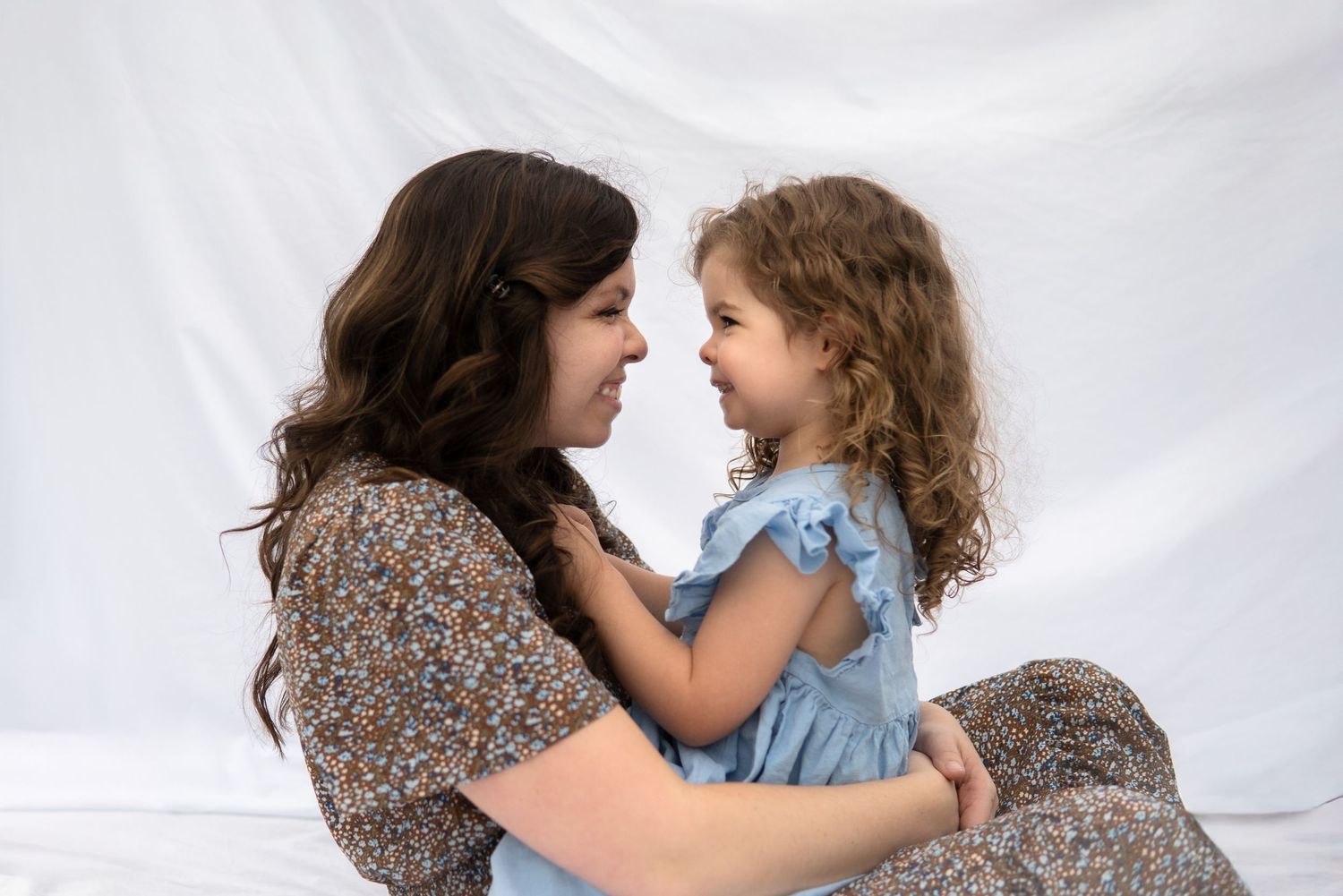 A parent and young child in blue dress share an intimate moment against a white backdrop.