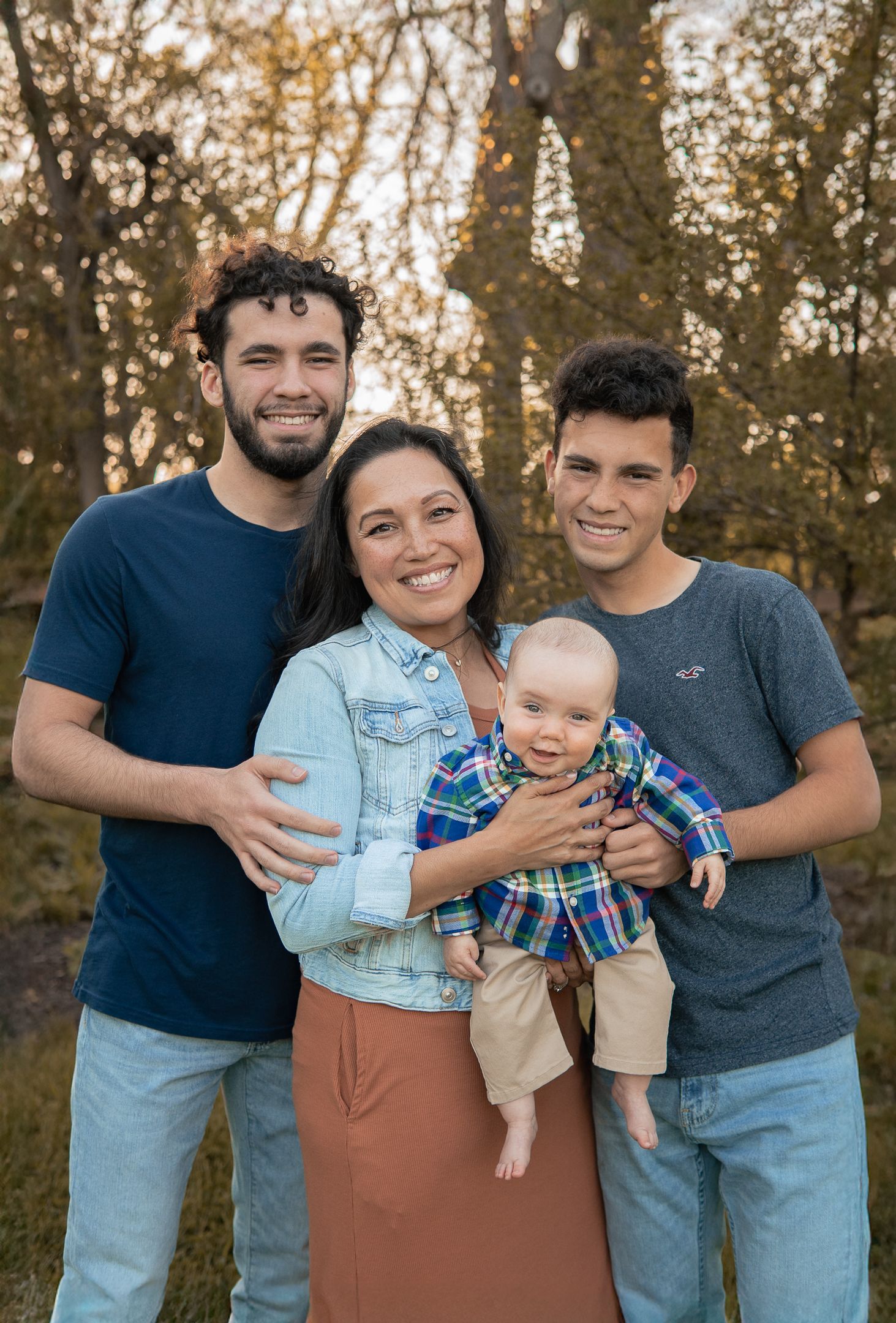 A family group poses together outdoors in casual attire during golden hour with autumn trees in the background.