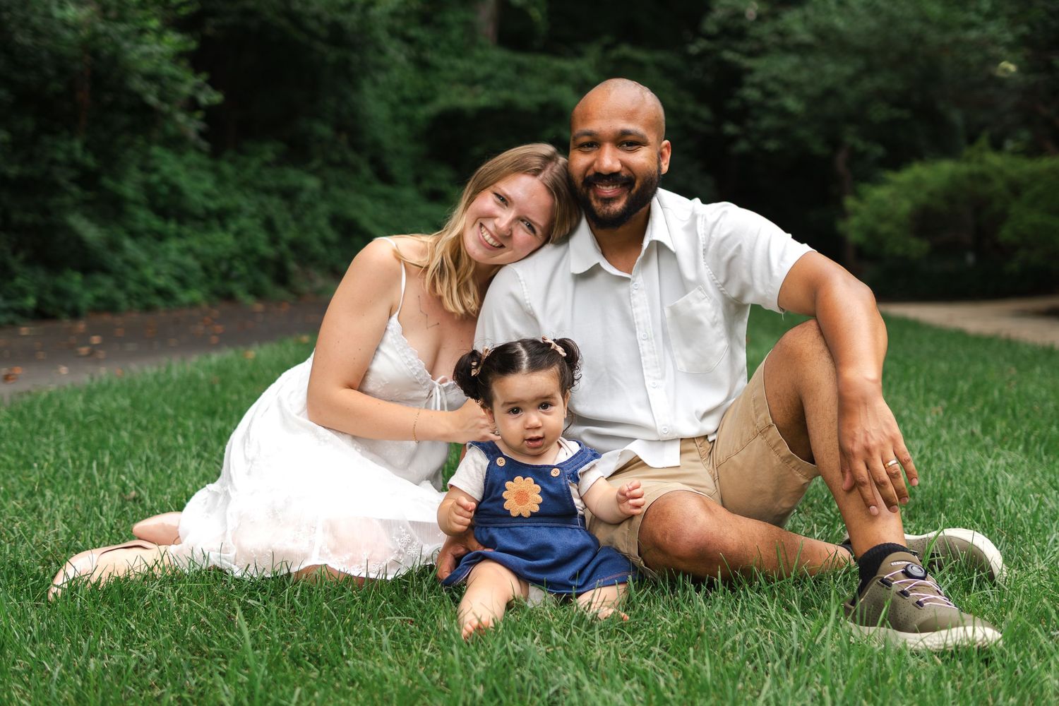 Family relaxes together on grass in outdoor portrait session at park, toddler sits in front wearing blue overalls.
