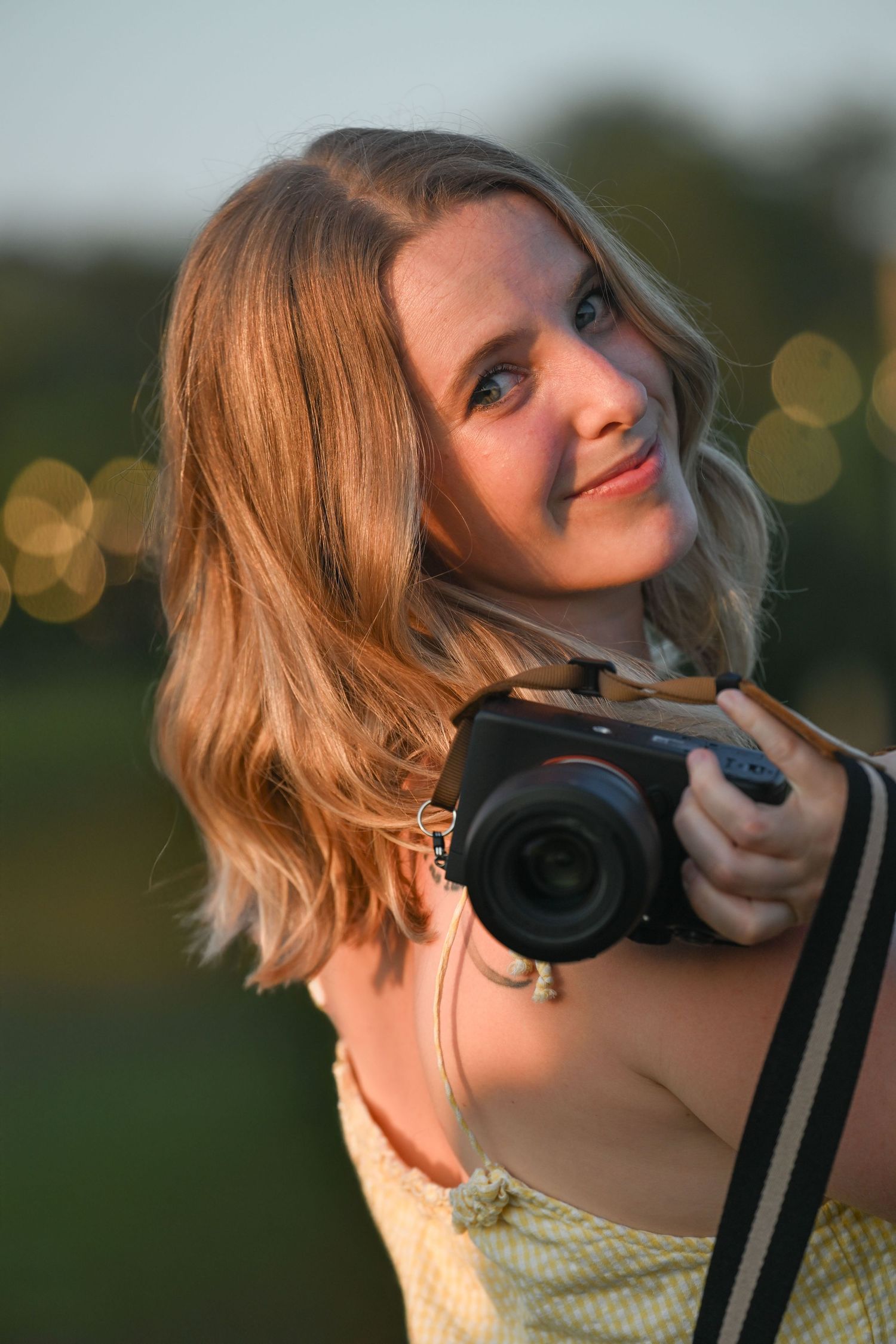 A photographer with wavy hair holds a camera while wearing a yellow dress at sunset.