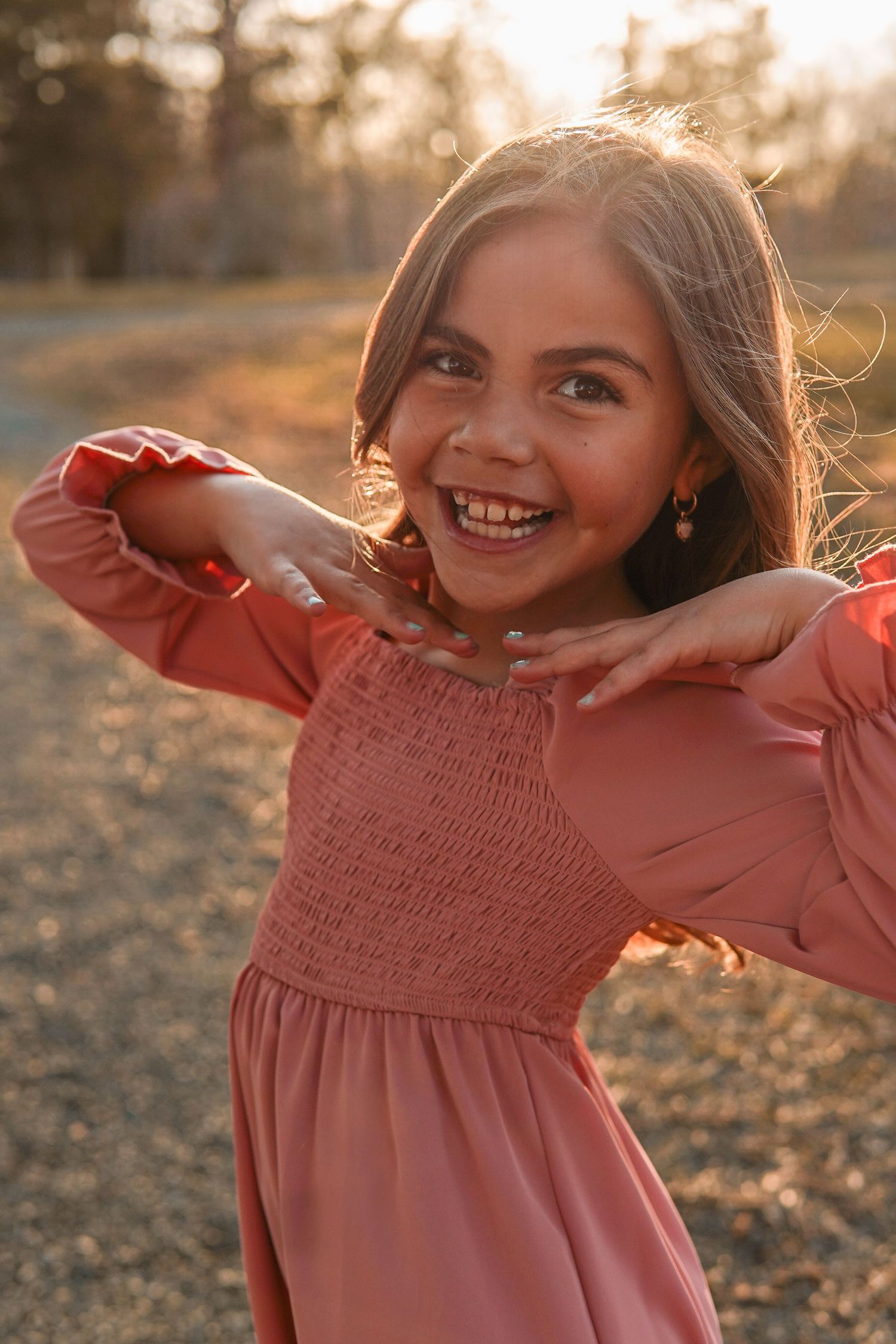 A child wearing a coral smocked dress smiles brightly in golden sunset lighting while playing outdoors.