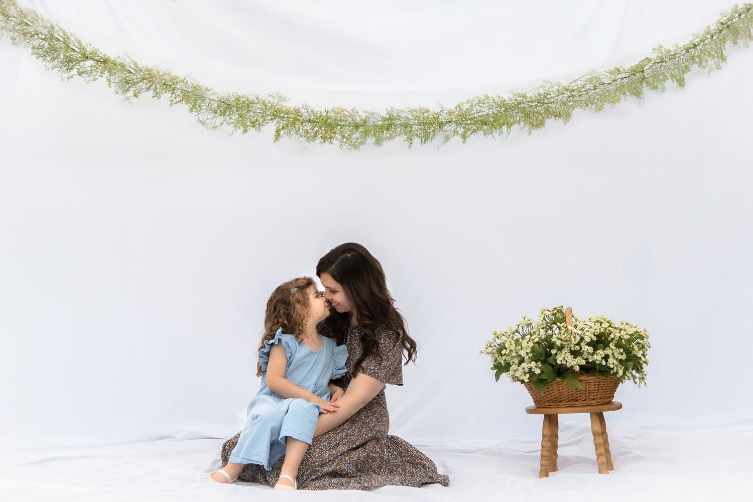 Two figures sitting together on a white floor under a delicate green garland with white flowers on wooden stool nearby.
