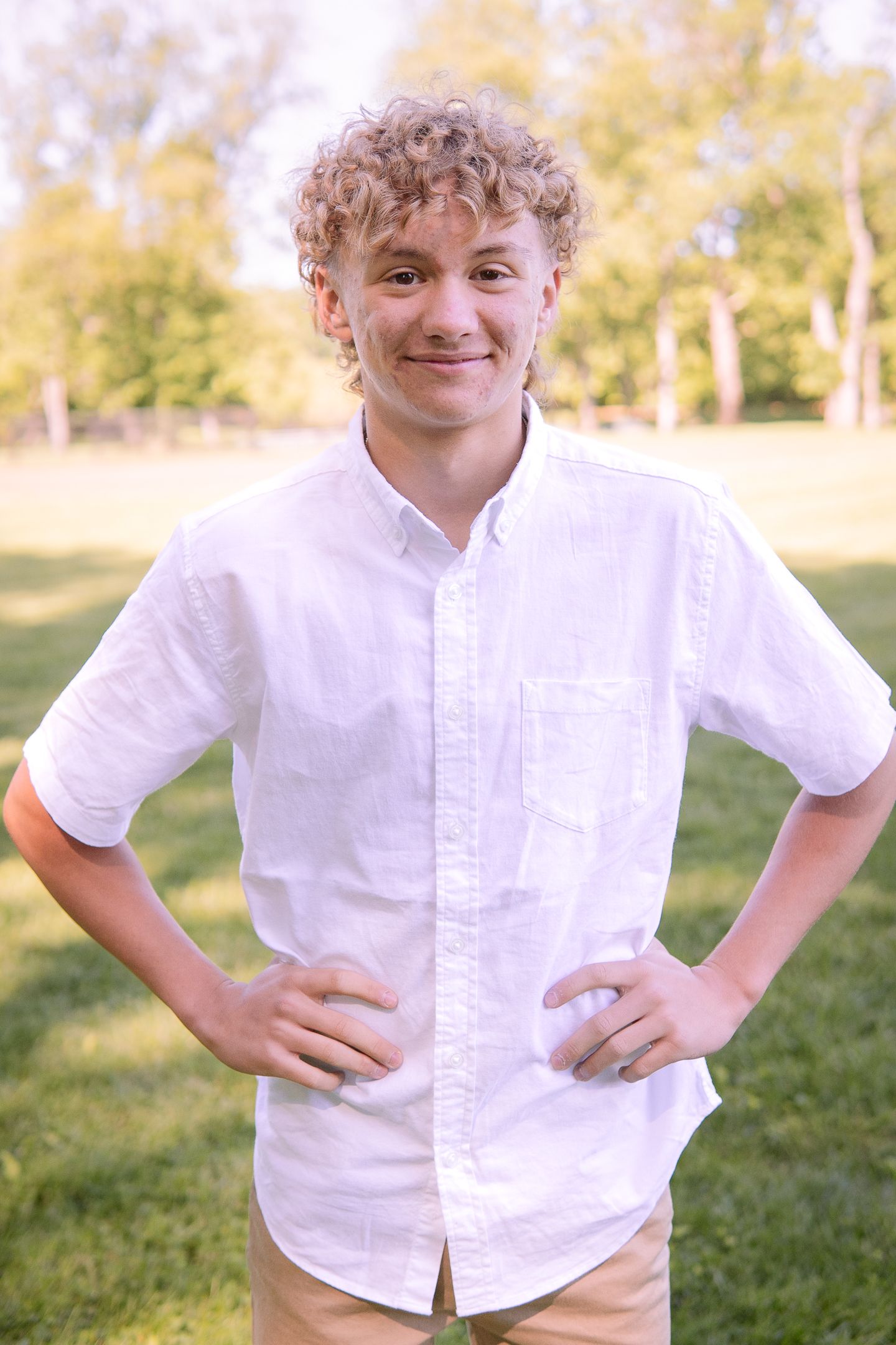 A teenager in a white button-down shirt poses confidently outdoors with hands on hips.