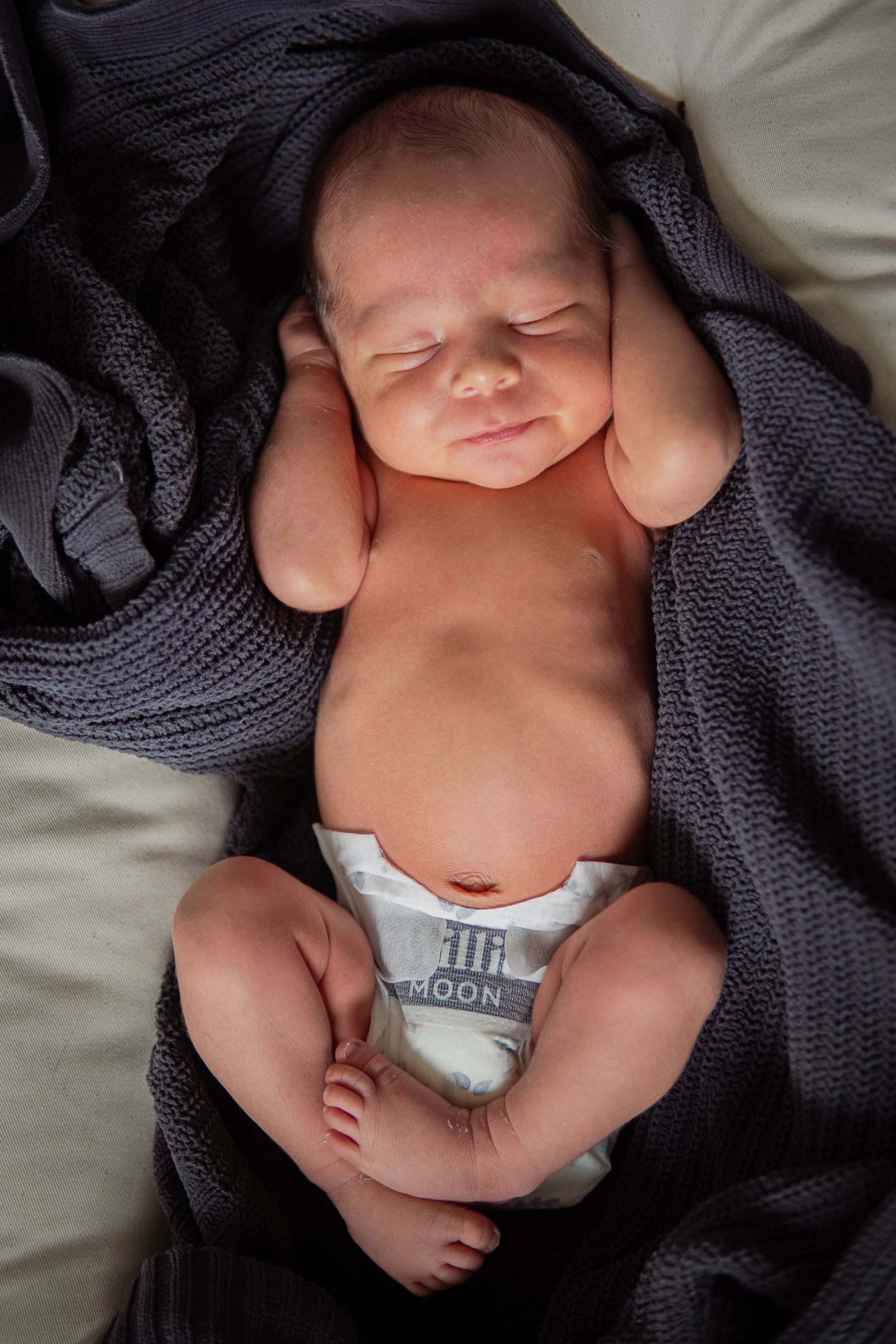 Newborn baby wrapped in dark knit blanket lying peacefully on white bedding.