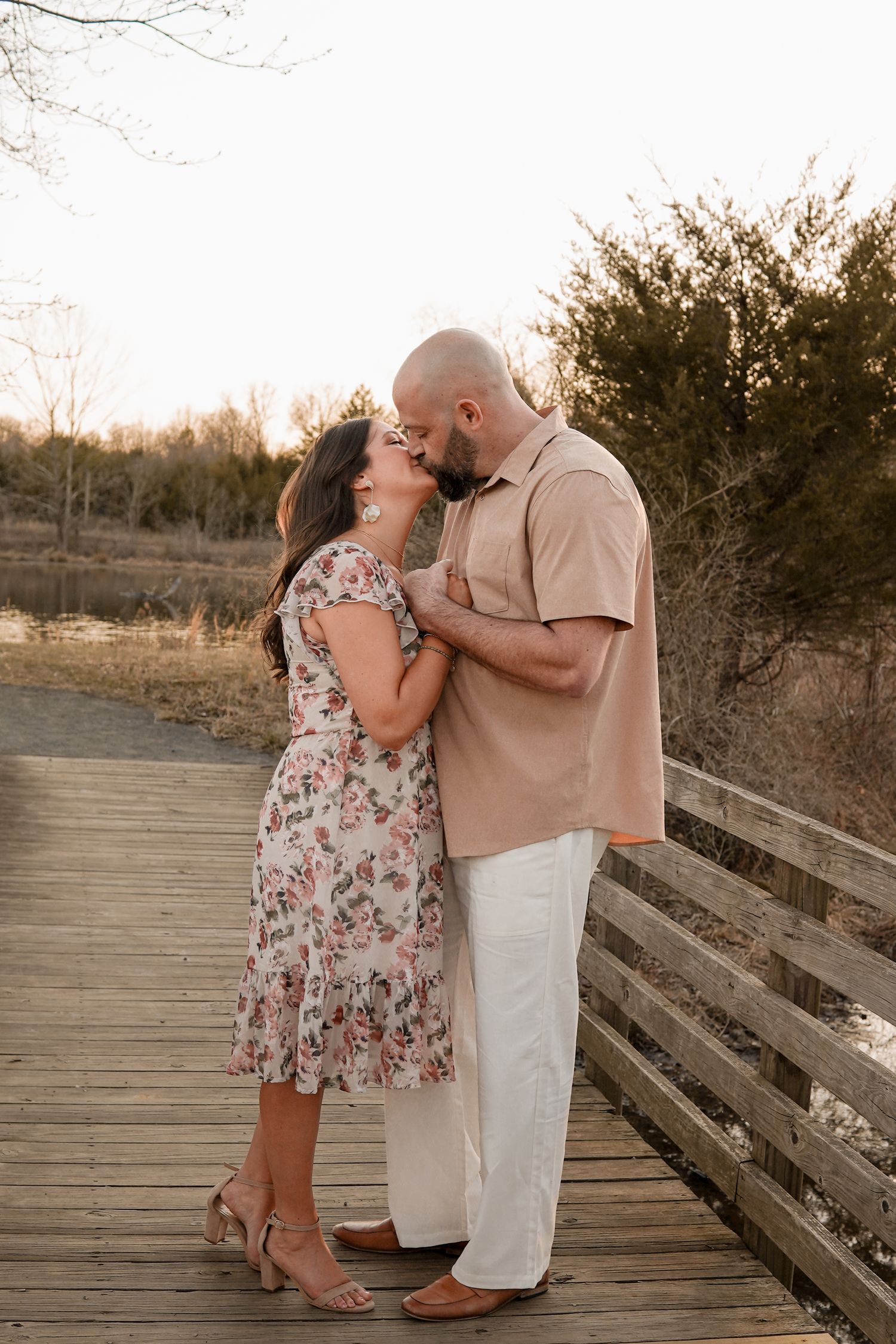 A couple shares romantic moments in floral dress and khaki attire on a wooden boardwalk during sunset.