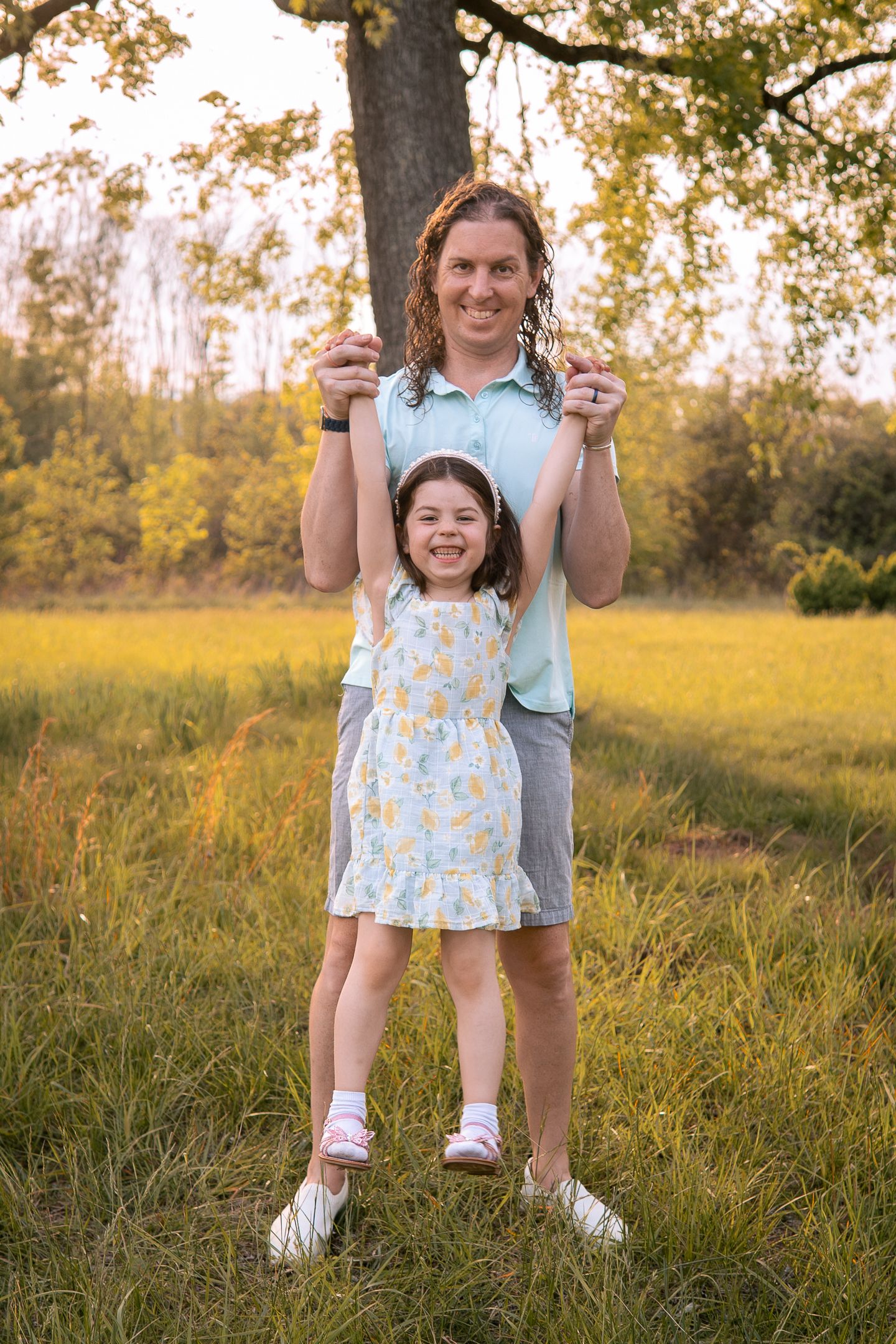 Two people stand together in a golden field during sunset with a large tree in the background.
