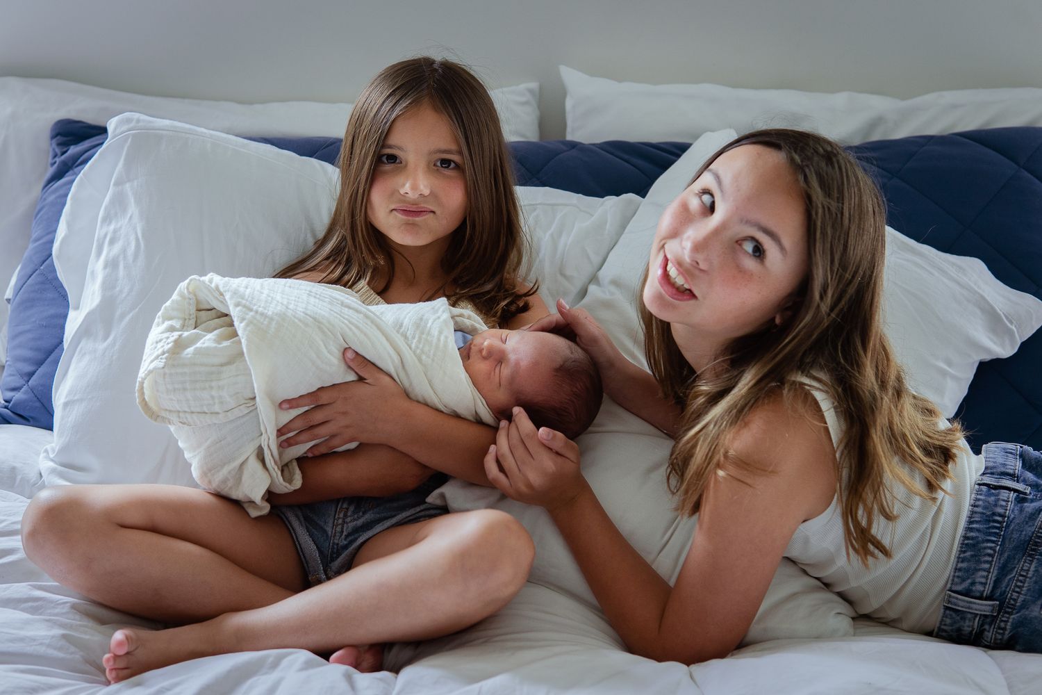 Young girls cuddling with newborn sibling on bed with white and blue bedding.