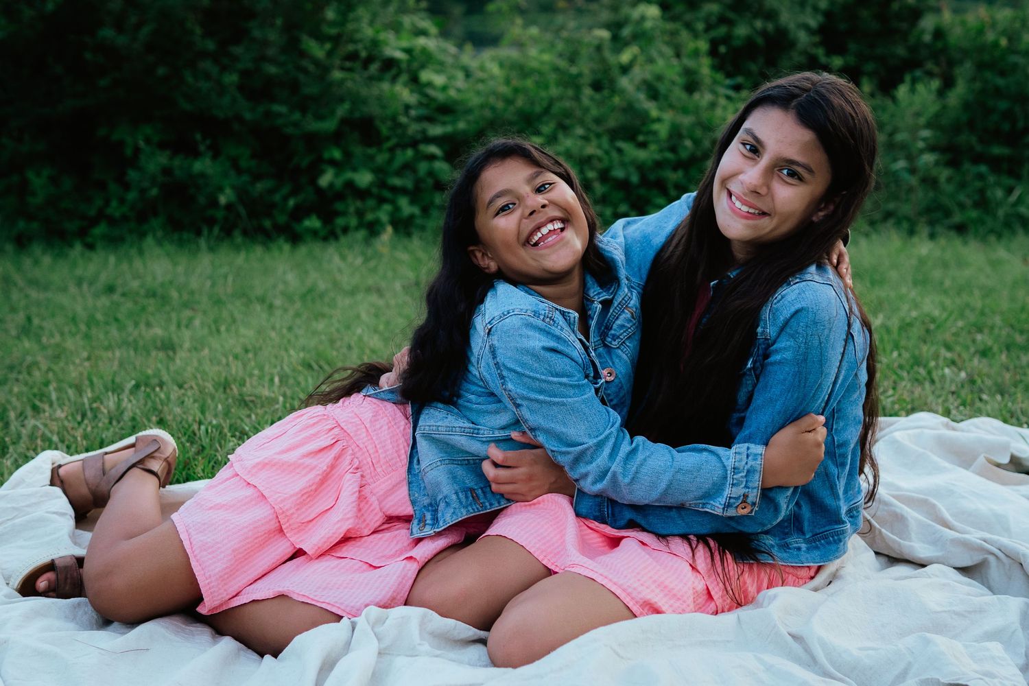 Two people lie on a white blanket outdoors, embracing and laughing together while wearing matching denim jackets.