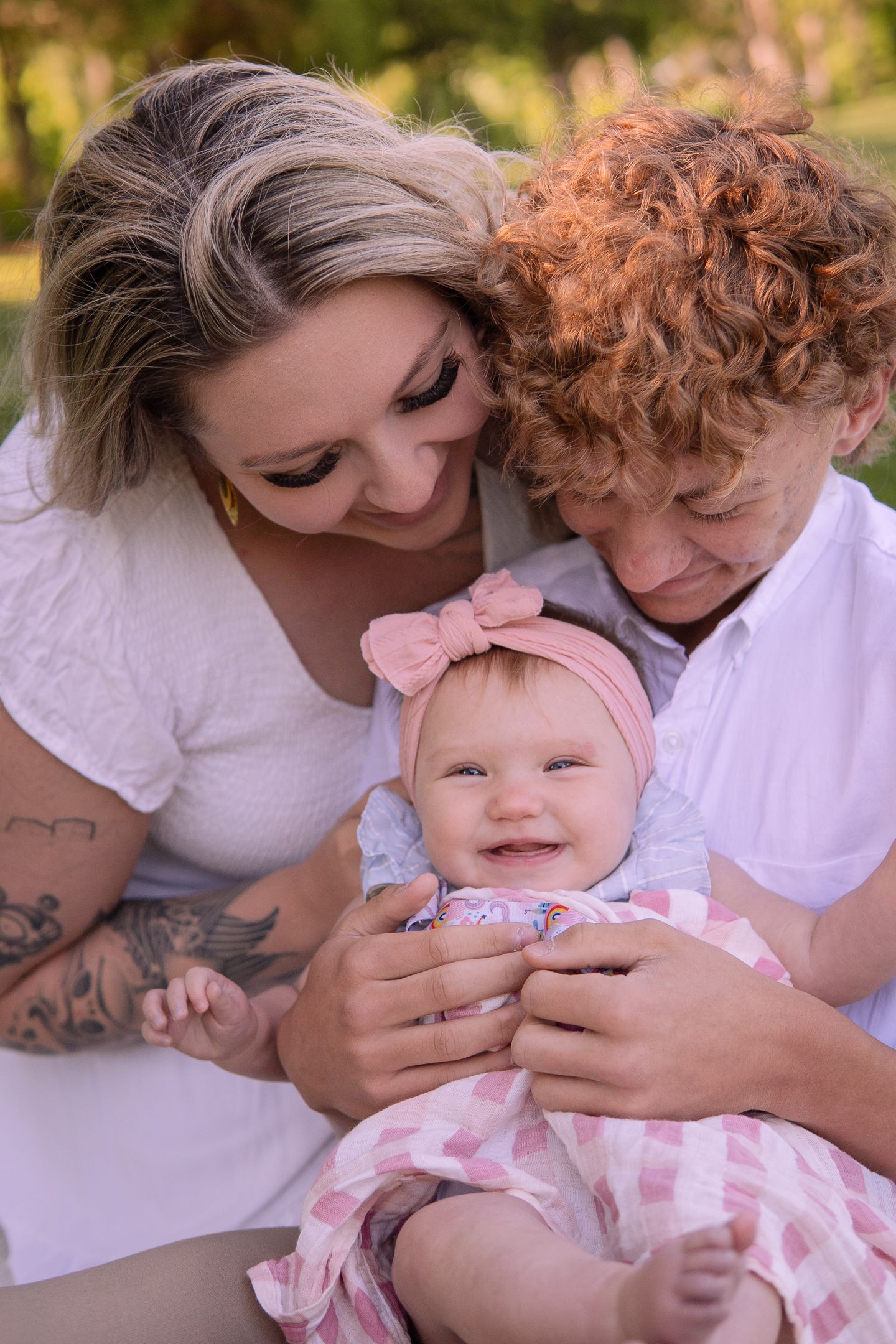 A close-up outdoor family photo with a happy baby in pink wearing a bow headband.