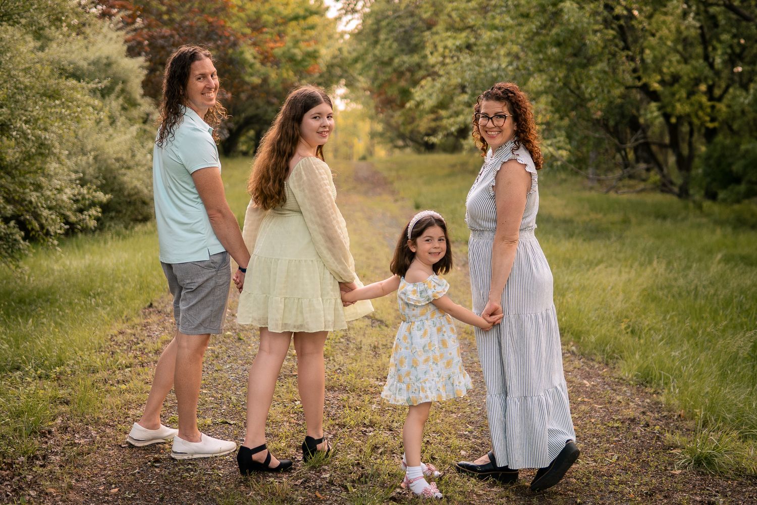 Four people stand together holding hands on a grassy path during golden hour in a natural outdoor setting.