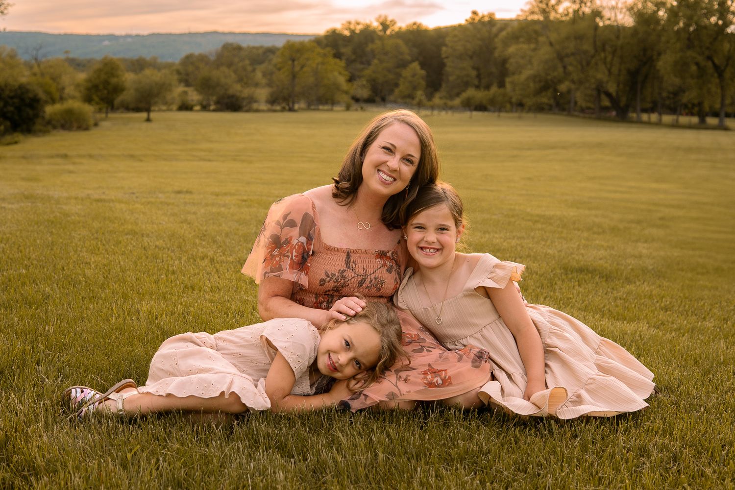 A family in coordinating pink outfits sits together on grassy lawn at sunset with rolling hills in background.