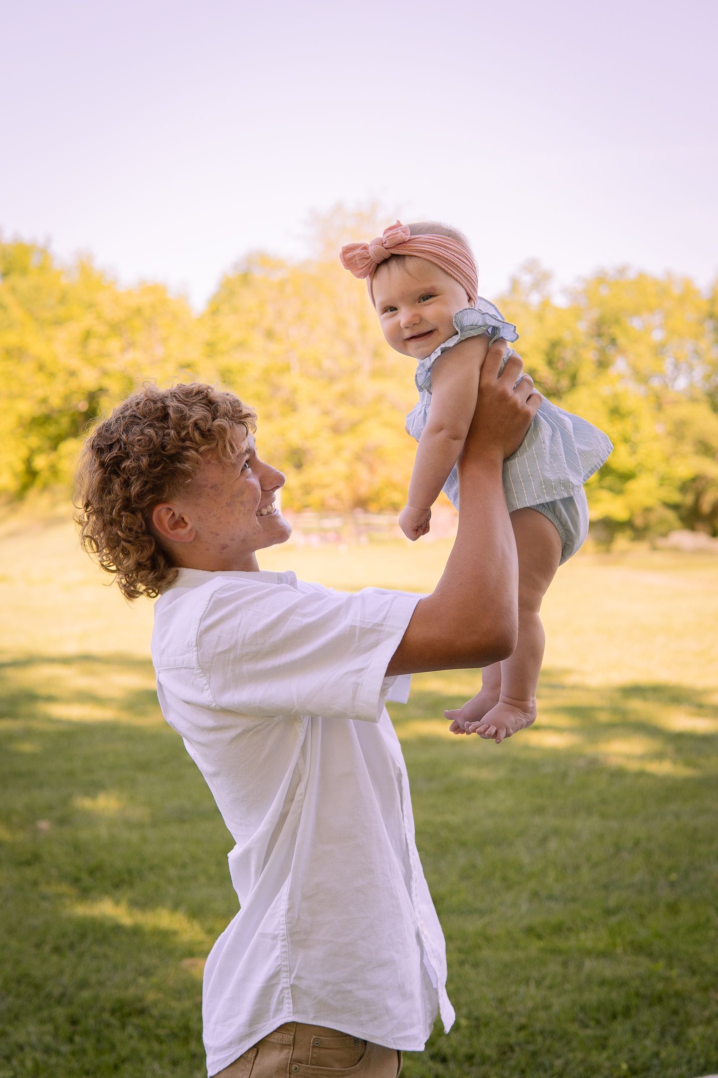 Parent playfully lifts a small baby in the air against a backdrop of autumn trees and golden sunlight.