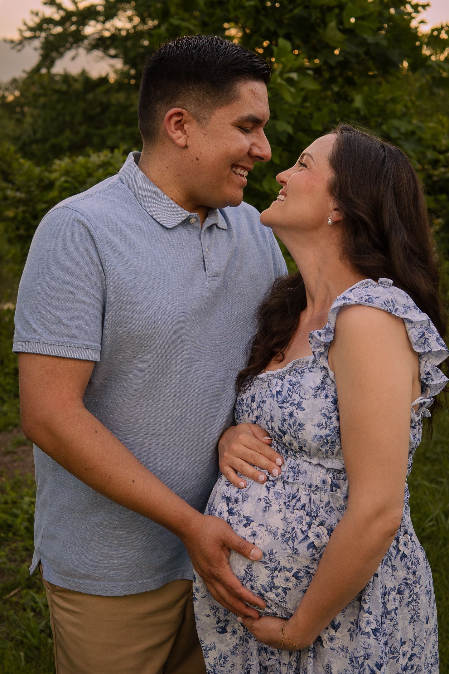 A couple shares an intimate moment outdoors during a maternity photoshoot at sunset, wearing grey and floral attire.