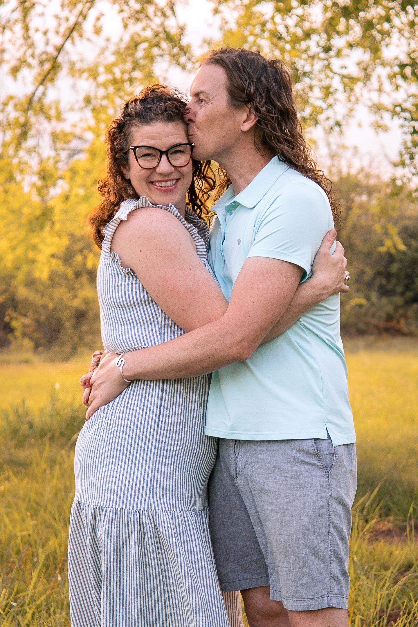 A loving couple embraces in a sunny field with golden grass and trees in the background during golden hour.