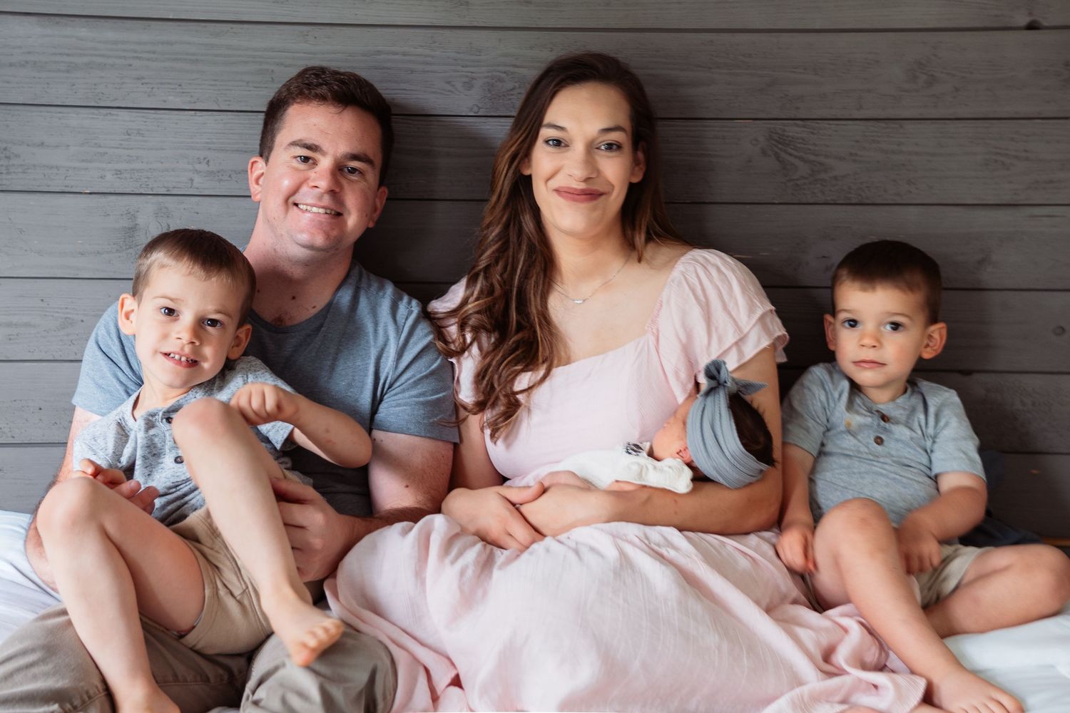 A family sits together against a gray wooden wall with their newborn.