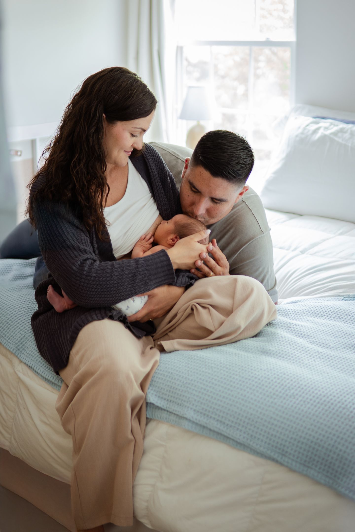 A couple sitting on a bed tenderly holds and gazes at their newborn baby in a sunlit bedroom.