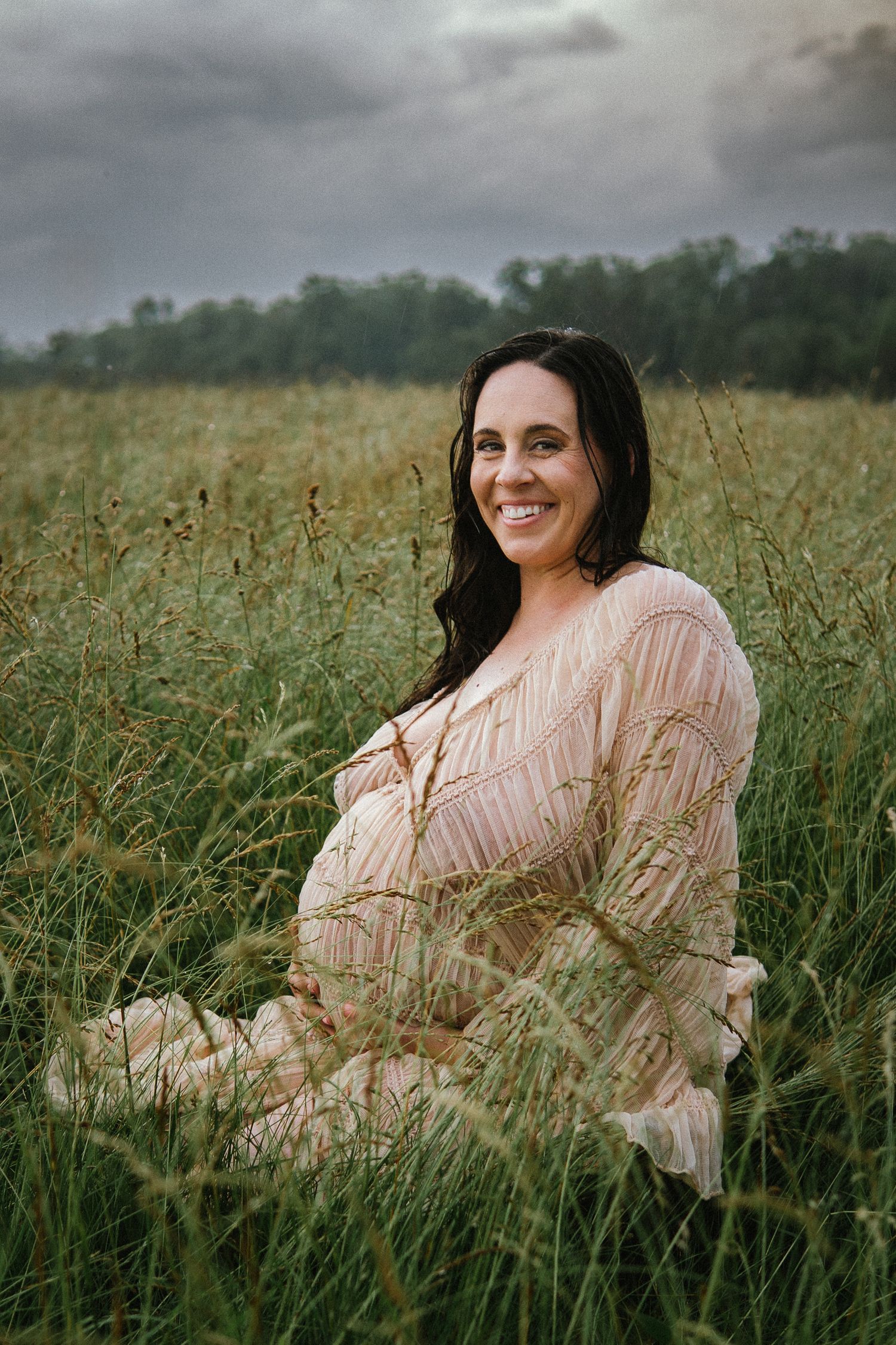 Expectant mother in flowing peach dress poses gracefully among tall grass in a dreamy outdoor setting.