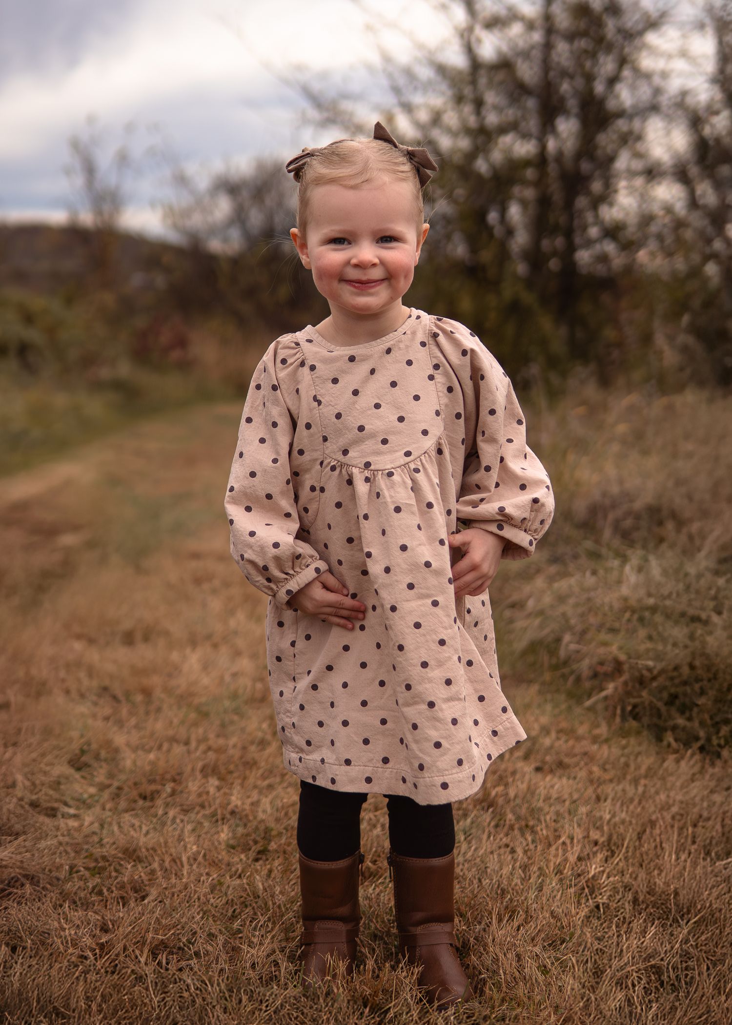 A child wearing a polka dot dress and brown boots stands in a grassy field on an autumn day.