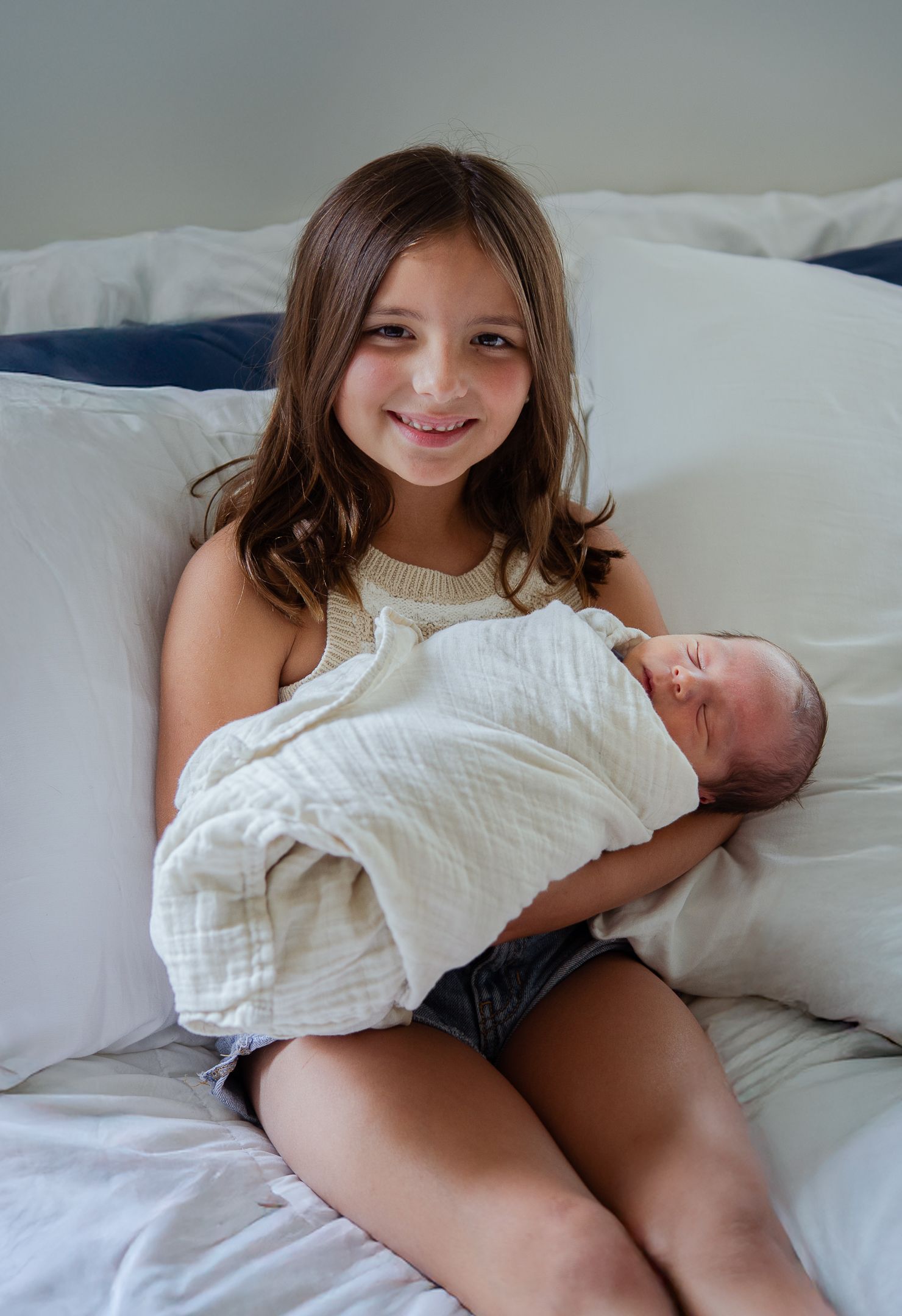 Series showing sibling bonding with newborn baby on white bed linens.