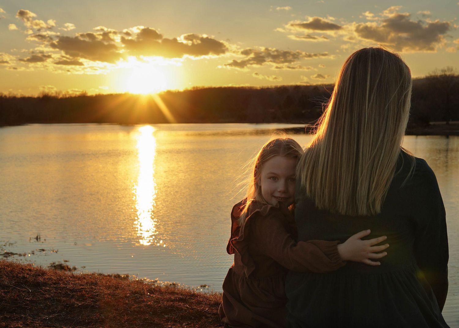 A silhouetted pair of people watching a golden sunset over a tranquil lake with sunbeams breaking through clouds.