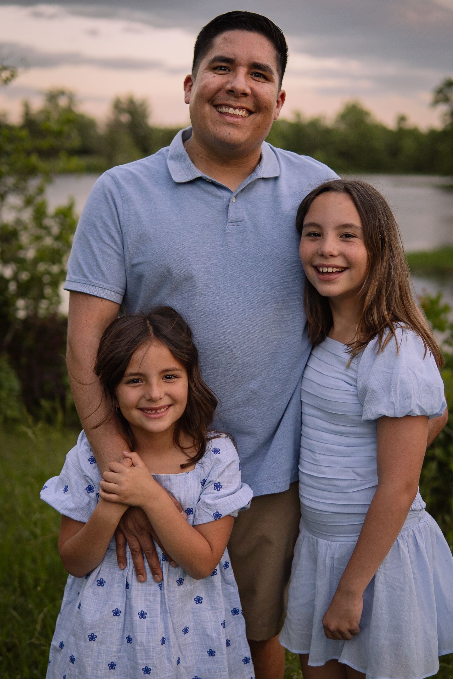 Family portrait with matching blue and white outfits standing by a lake at sunset.