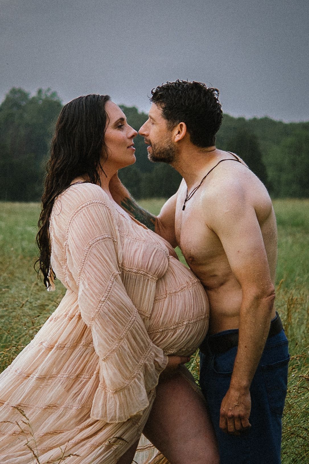 A romantic couple embraces in a field wearing pink and denim while creating a dreamy maternity photo series.