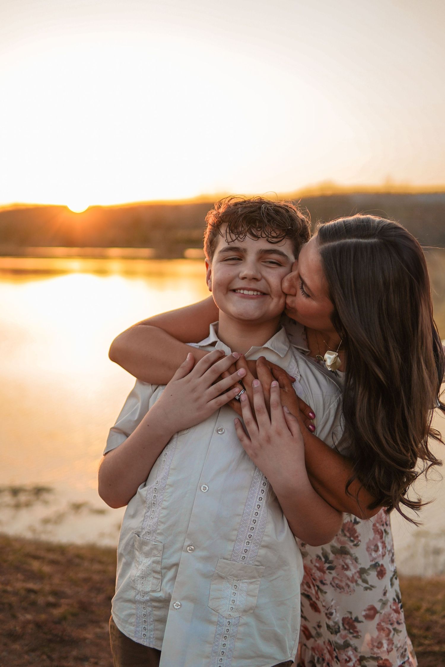 A loving embrace between siblings captured at sunset by a lake with warm golden light glowing in the background.