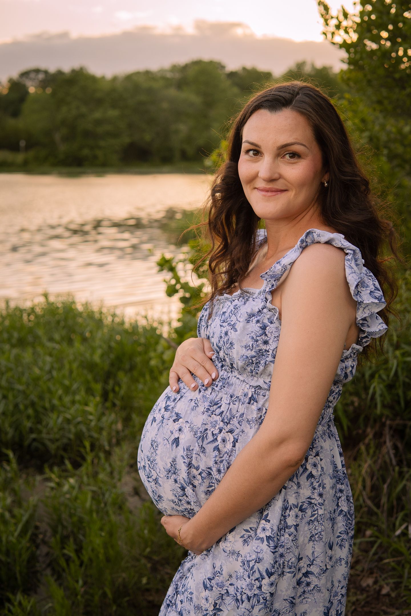 A pregnant woman in a floral blue dress stands by a lake at sunset during a maternity photoshoot.