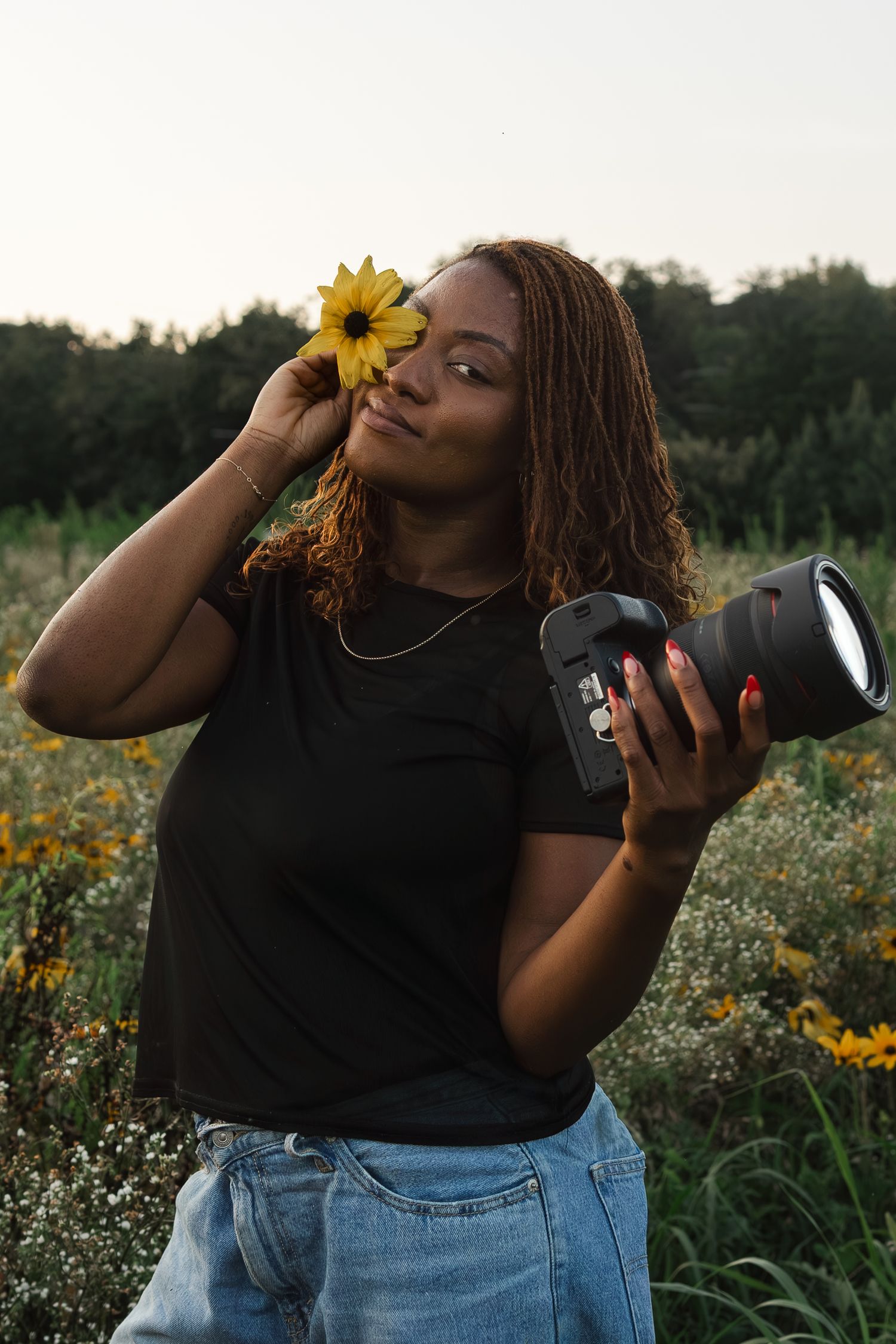 Creative photographer holds camera and sunflower while posing in wildflower field at sunset.