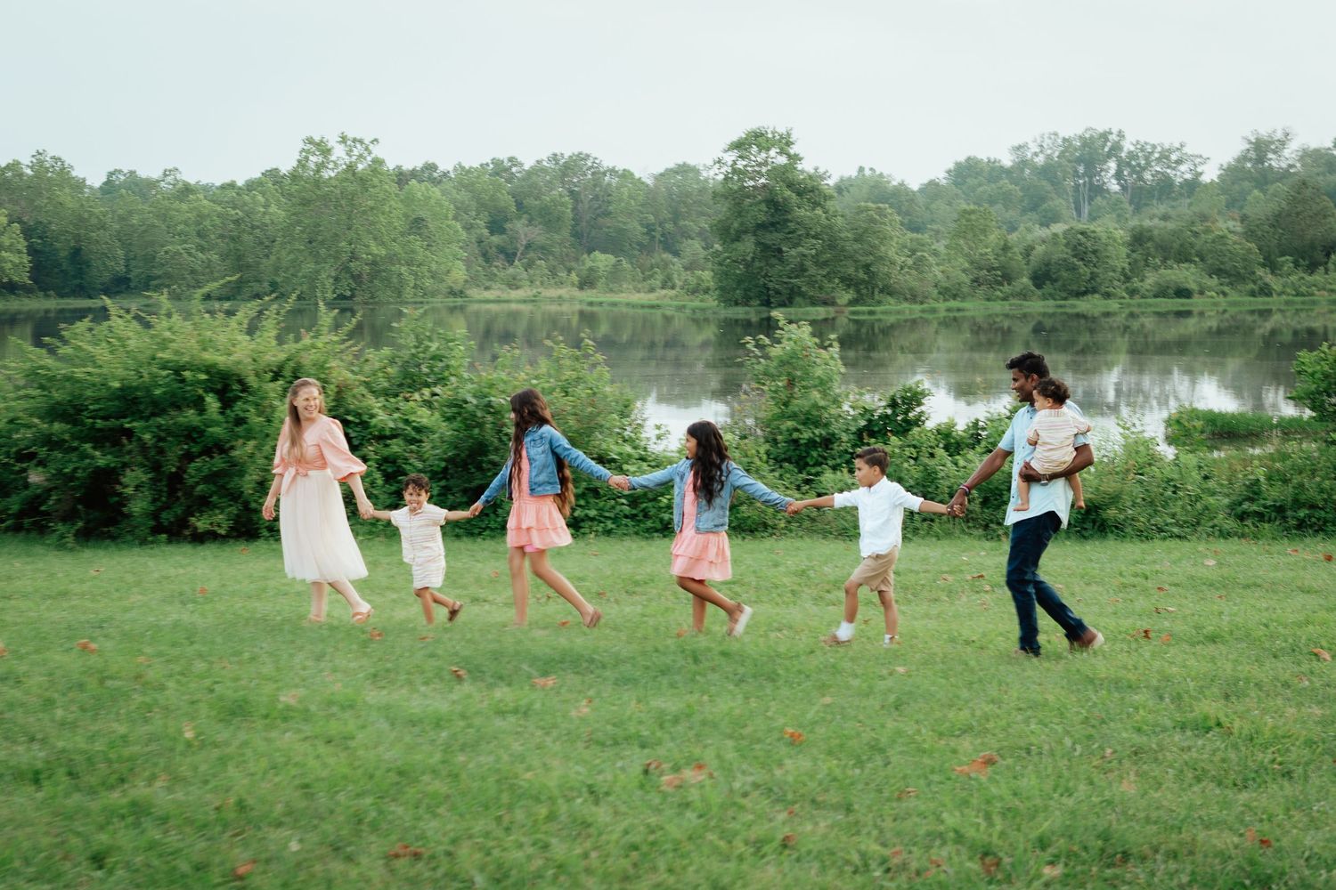 A sequence of people holding hands and dancing in a line across a green meadow on a sunny day.