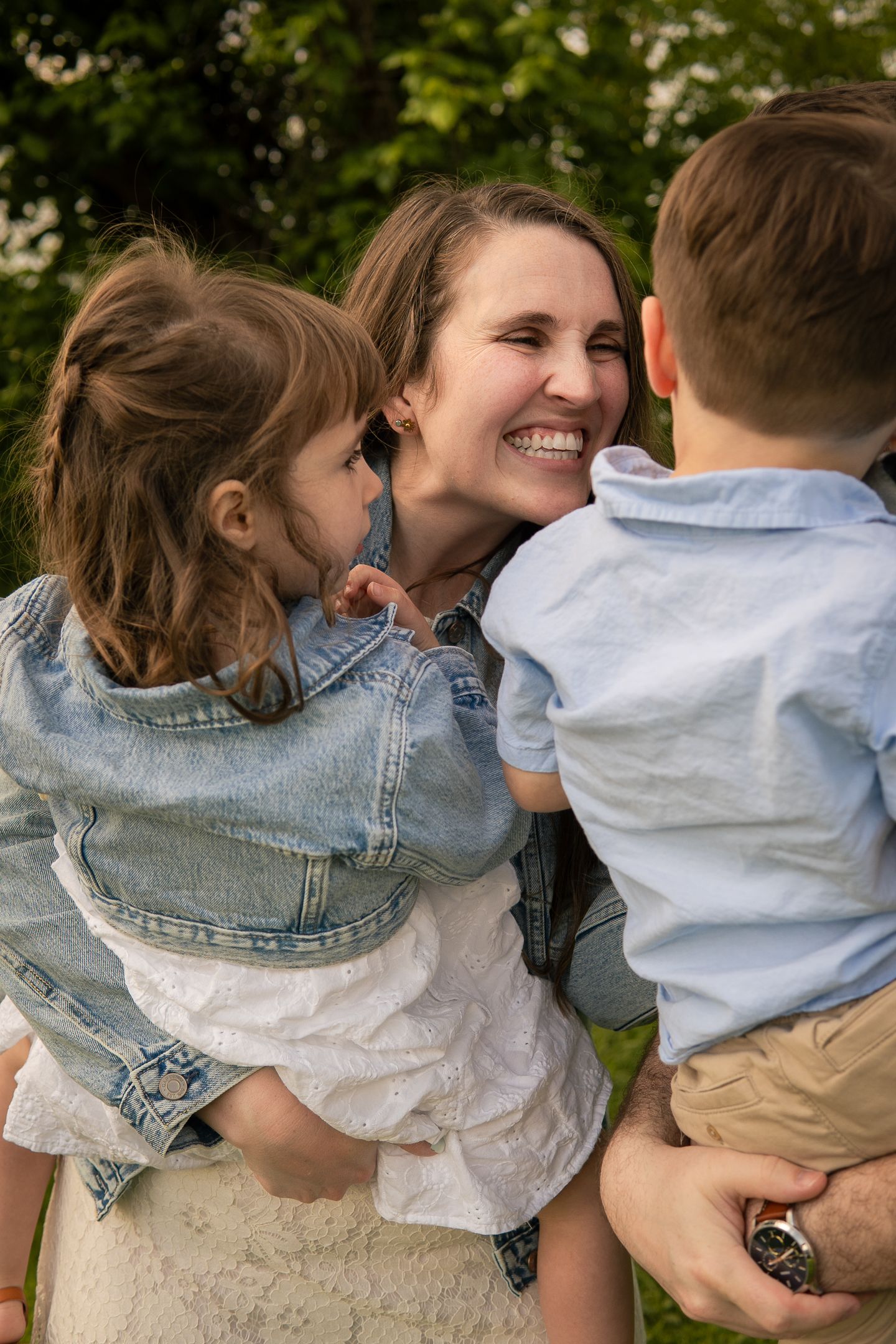 A joyful outdoor family moment showing a woman holding two children in casual denim and light colored clothing.