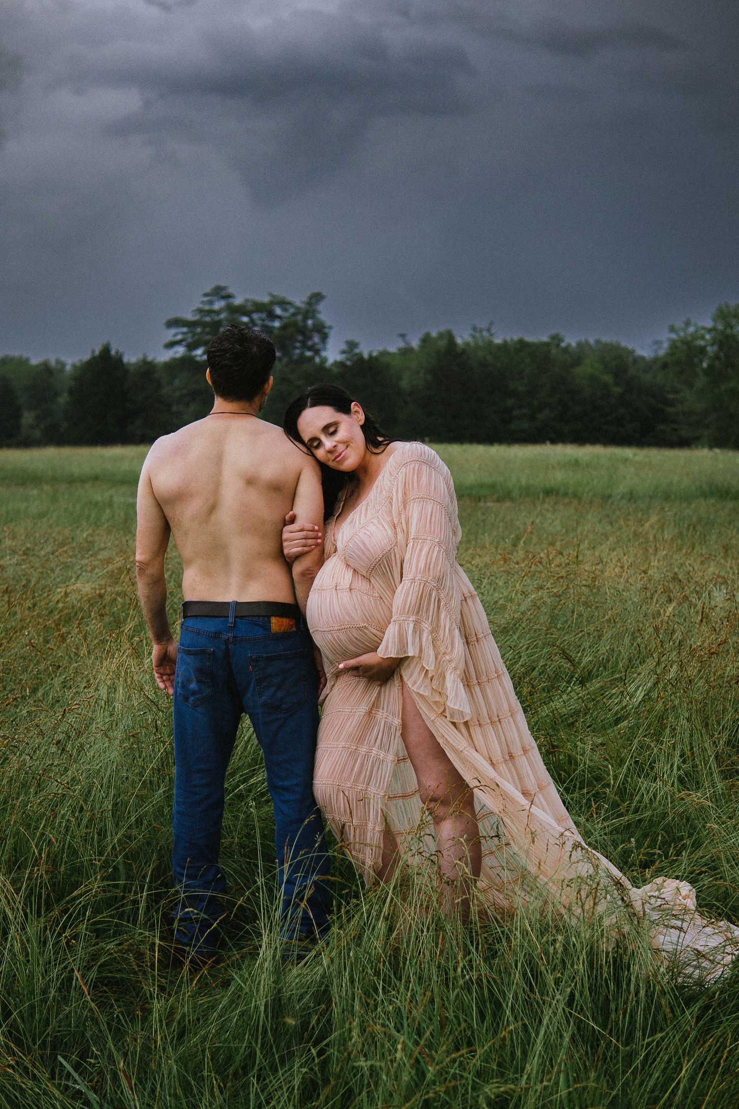 A maternity photo in a field shows couple embracing under dramatic stormy skies.