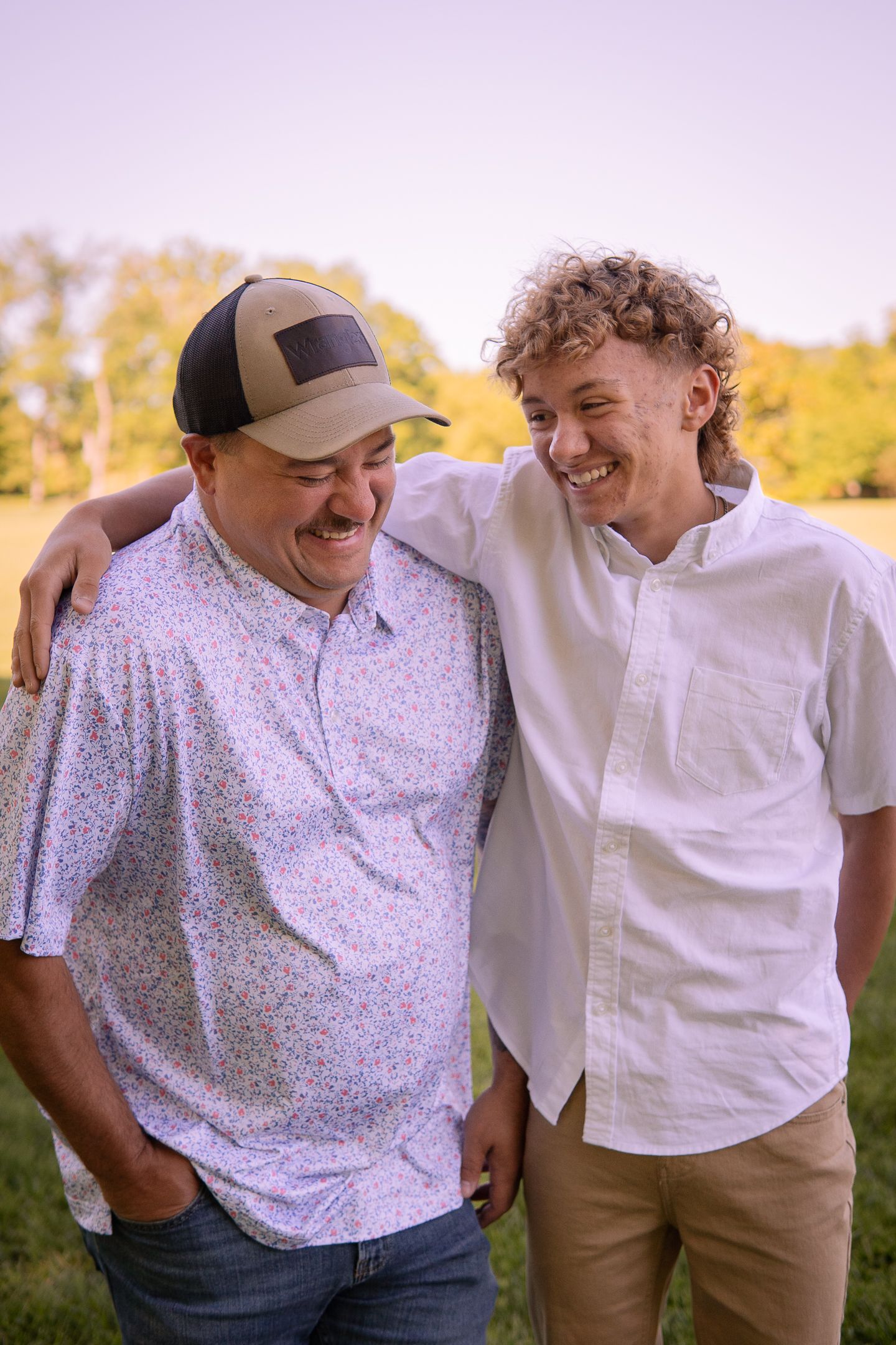 Two figures share a candid laughing moment outdoors wearing casual attire against autumn foliage.