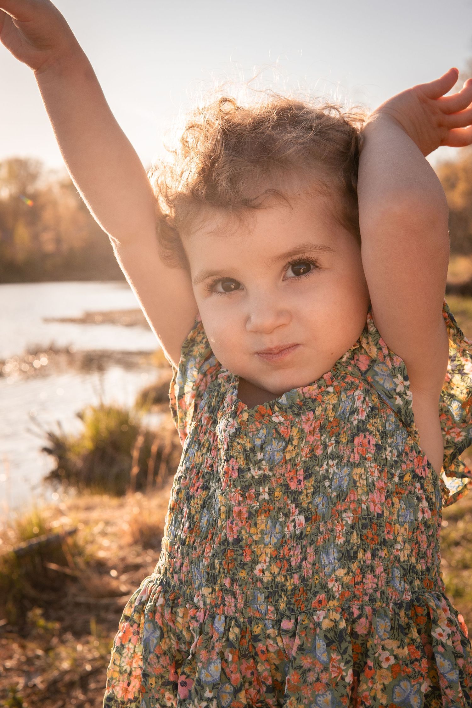 A young child in a floral dress stands outdoors in golden sunlight beside a lake with arms raised joyfully in Haymarket, Virginia.