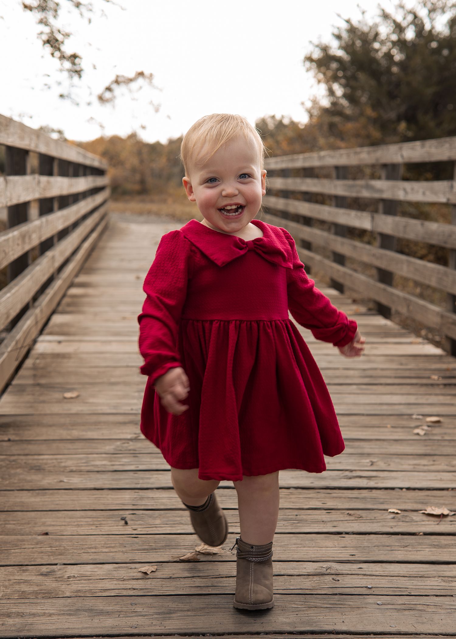 A small child in a red velvet dress and brown boots walks along a wooden boardwalk on an autumn day.
