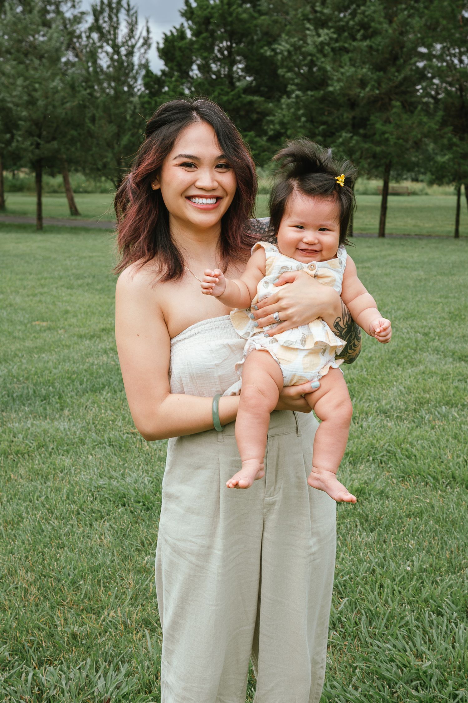 A candid outdoor portrait of a parent in a beige dress holding a smiling baby in a floral outfit on a grassy lawn.