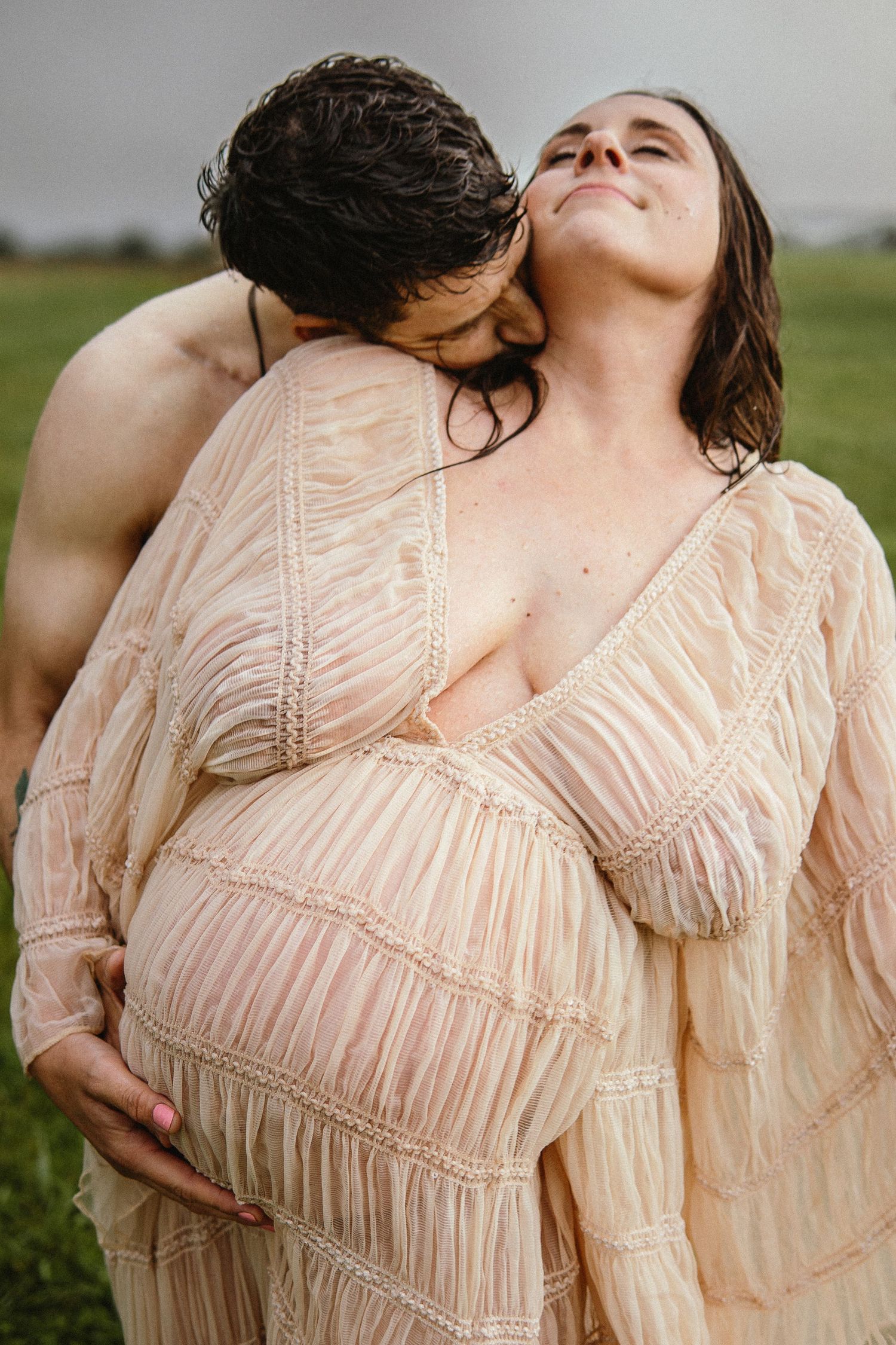 A romantic maternity photoshoot in a field shows an intimate moment in a flowing beige dress.