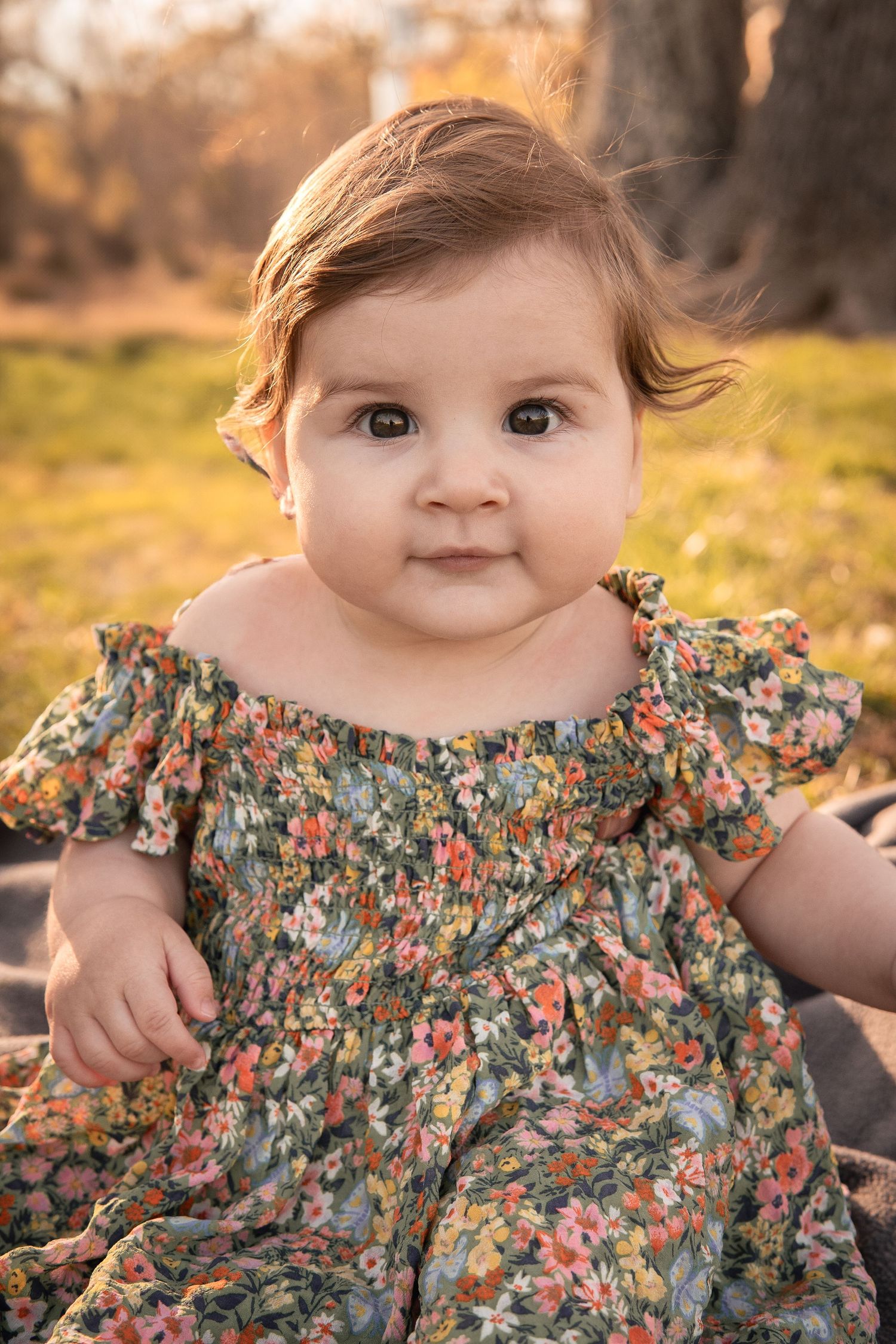 Baby in floral print dress sits outdoors during golden hour sunset with autumn trees in background in Haymarket, Virginia.